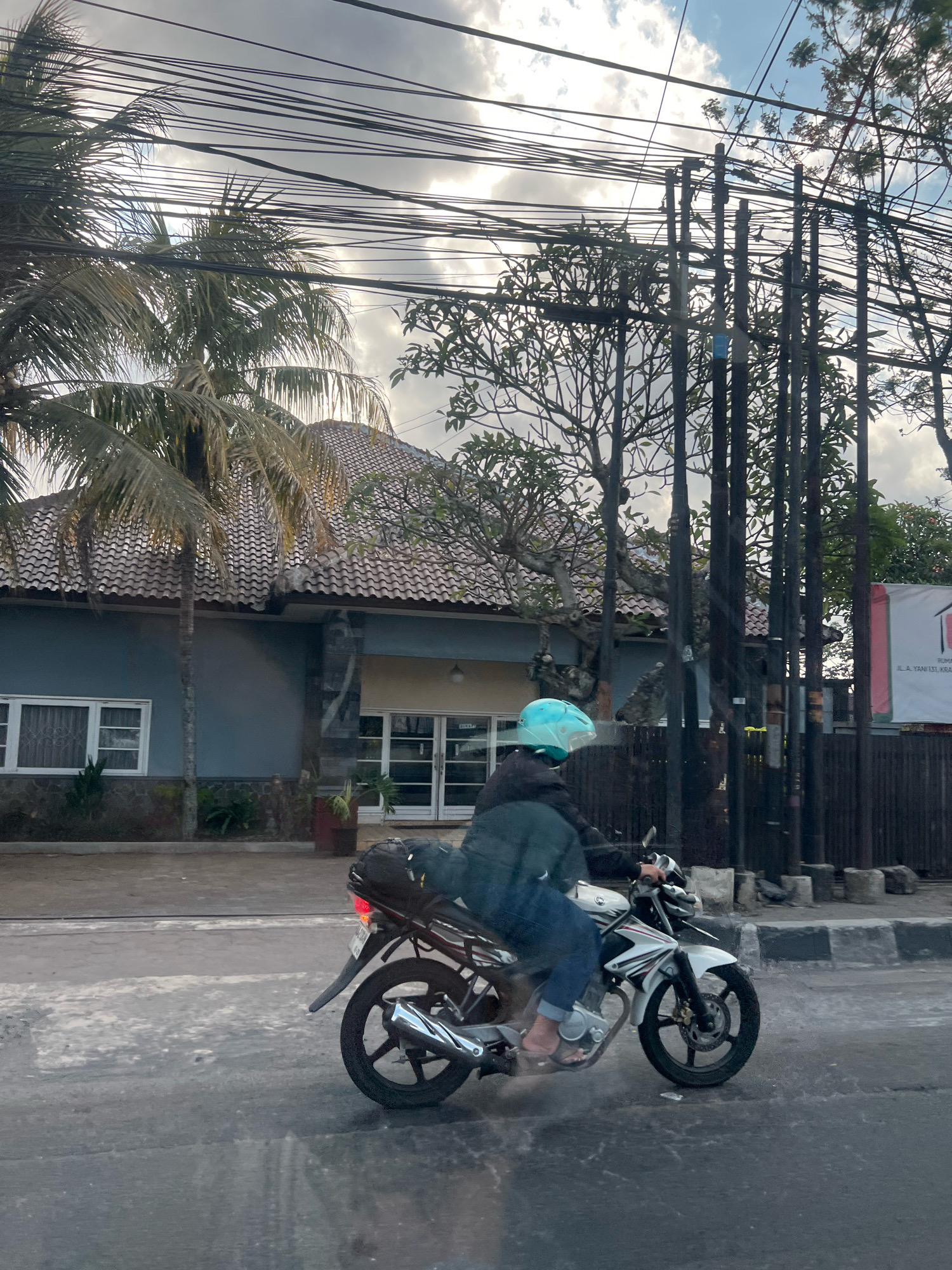 A person wearing a blue helmet and a dark jacket rides a white motorcycle with a black bag on the back. The motorcycle is on a wet, paved road. Behind the motorcycle, there is a building with a tiled roof and several windows. Palm trees and other foliage are visible. Utility poles and wires run across the sky, which is cloudy. A banner is visible on the right side of the image. The scene appears to be in a residential or commercial area. The context of Magelang, Indonesia, suggests a tropical or subtropical environment, which is consistent with the presence of palm trees and the style of the building.