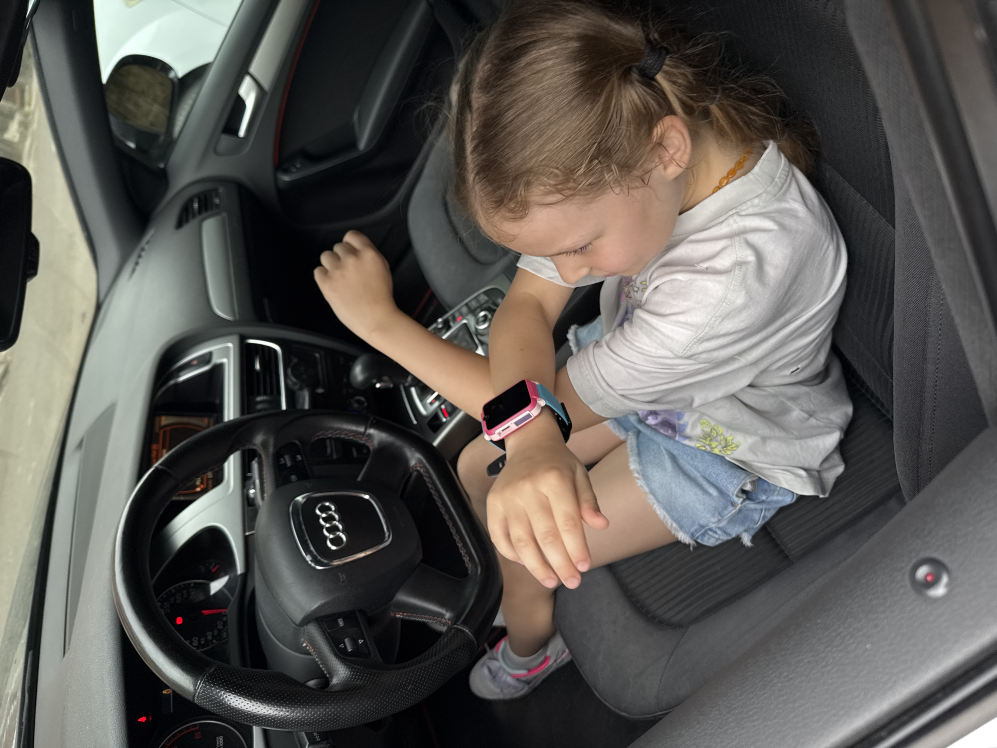 A young girl sits in the driver's seat of an Audi sedan. She wears a white t-shirt, denim shorts, a necklace of amber-colored beads, and a pink and blue smartwatch. The car's dashboard and steering wheel are visible. The location is Budva, Montenegro.