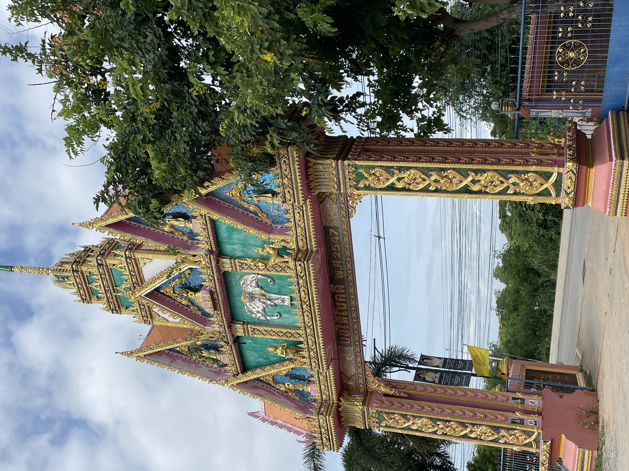 The image shows an ornate, multi-tiered gateway with a pink and gold color scheme, decorated with intricate patterns and sculptures. At the top of the gateway, there is a spire. The middle section of the gateway features a turquoise background with a silver elephant sculpture and smaller figures. Below this, there is text in Thai script. The gateway is flanked by large pillars adorned with gold detailing. To the left of the gateway, there are two black banners hanging, one of which displays portraits. A yellow flag is also visible. The gateway opens onto a road, with dense green foliage and trees visible on either side, and a blue metal gate to the right. In the background, there are power lines and a cloudy sky. This gateway is identified as belonging to Wat Nong Yai Buddha (วัดหนองใหญ่) in Pattaya City, Thailand, and the text on the gateway appears to be a date in the Buddhist Era, "พ.ศ. 2547" which translates to 2004 CE.