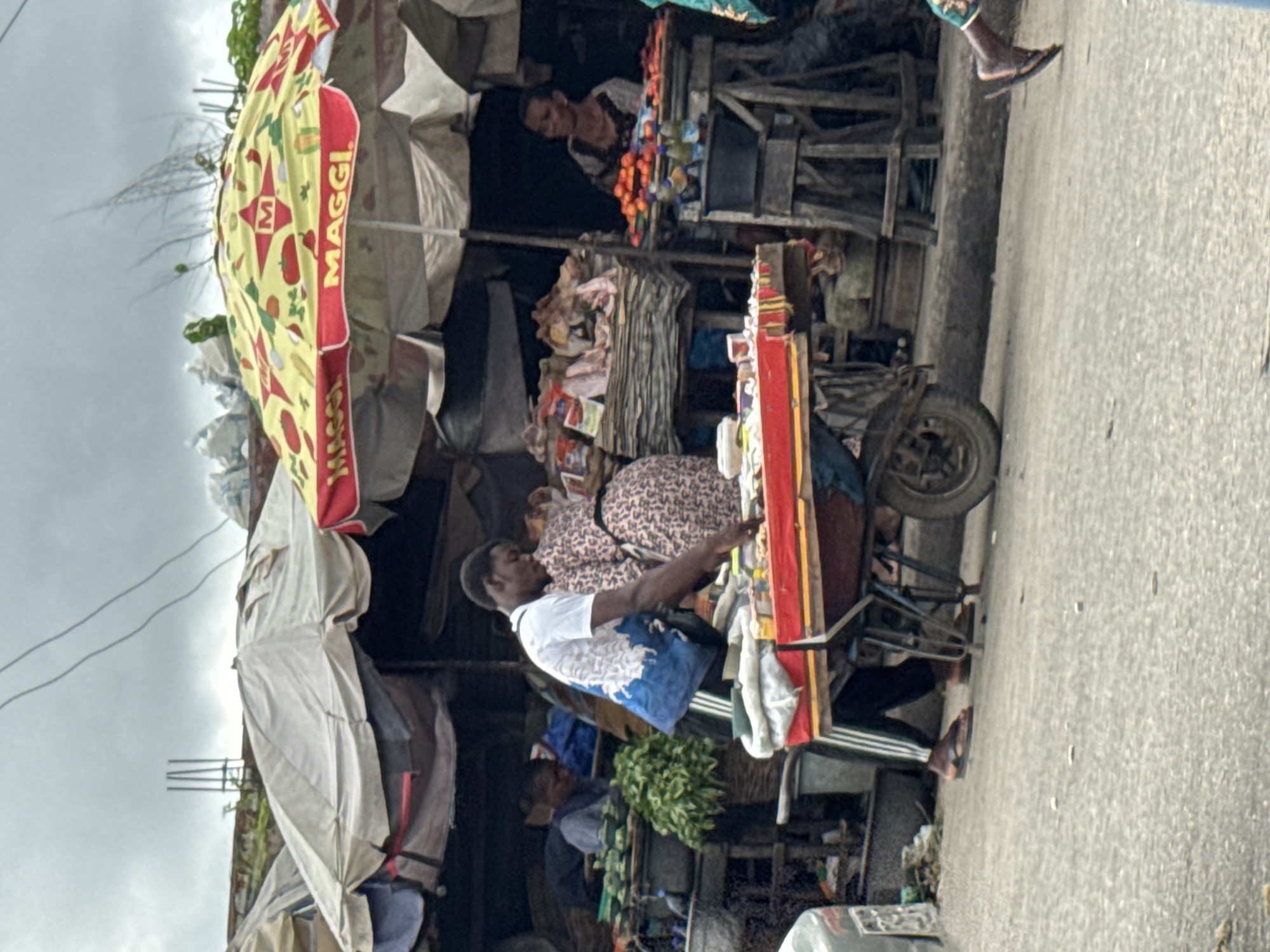The image depicts an outdoor market scene in Badagry, Nigeria. A man in a white t-shirt and blue patterned bag is interacting with a woman wearing a leopard-print dress. They are positioned near a red and yellow cart, likely a vendor's stall, with various items displayed. The cart itself appears to be part of a larger market setup, with tents and structures visible in the background. The lighting suggests it is daytime, with overcast skies. The ground is paved, indicating a road or walkway within the market. The scene suggests a typical commercial activity, with people engaged in transactions or perhaps a brief interaction at the market. The Maggi logo is visible on a yellow umbrella, providing a brand context.