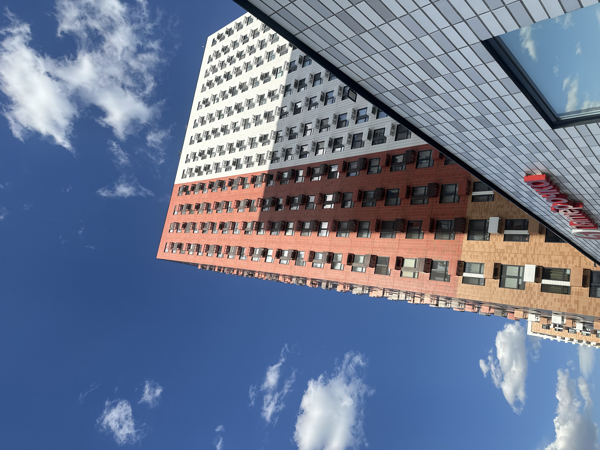 The image shows a tall, modern residential building in Lyubertsy, Russia. The building is divided into sections with different colors and textures, with white, red, and brown sections. Many of the windows are open. The building is set against a clear blue sky with scattered white clouds. A part of another building is seen at the top right, with a roof structure and a red sign.