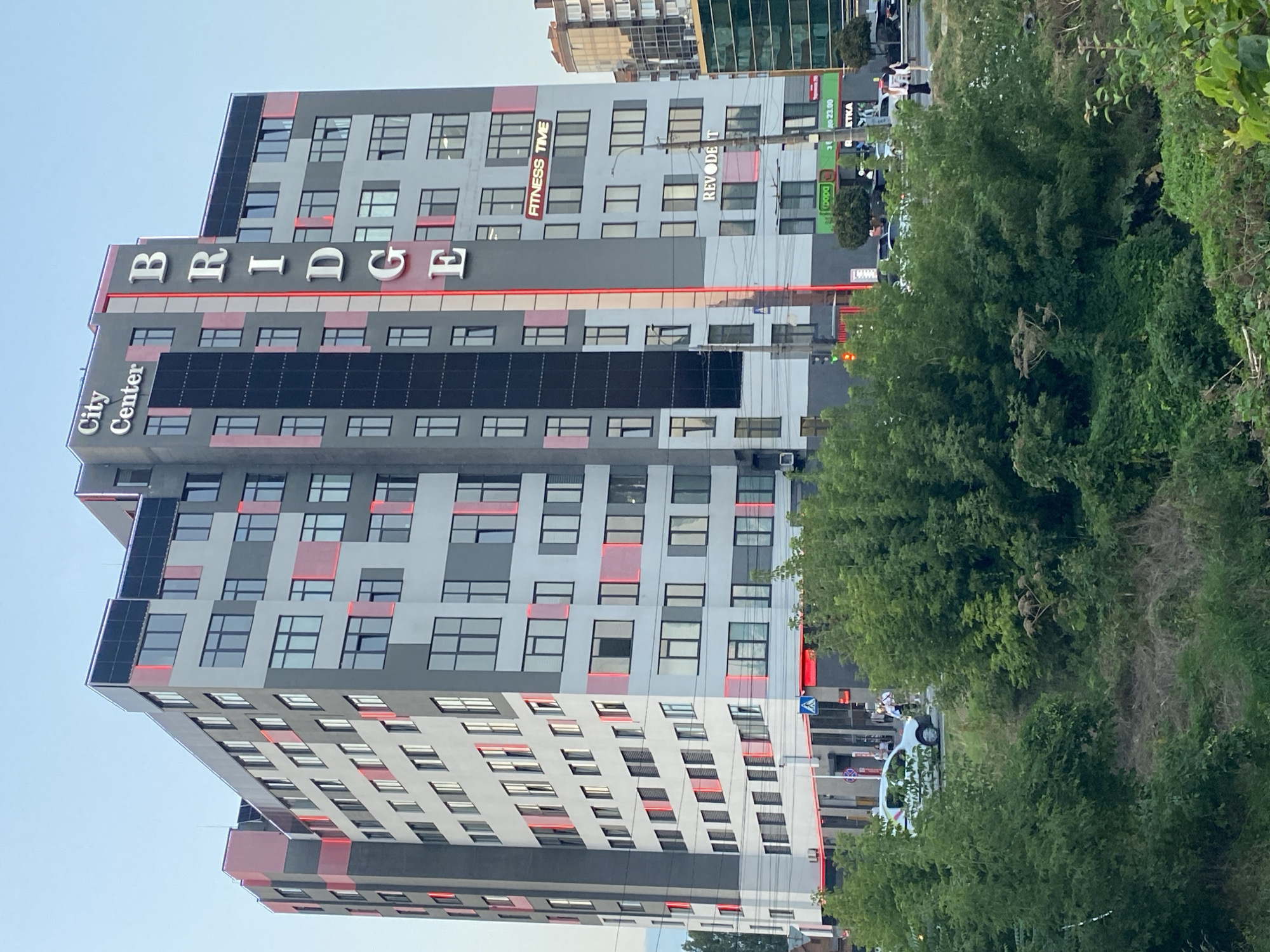 The image shows a multi-story building located in Vinnytsia, Ukraine, specifically on Soborna Street. The building, named "BRIDGE City Center," features a modern architectural design with a gray and light gray facade, accented by red lighting and dark panels. The name "BRIDGE" is prominently displayed vertically in large white letters on the side of the building, with "City Center" written above it. There is also a sign for "FITNESS TIME" and "REVODENT" on the building.
In front of the building, there is a street with a few cars, including a white car. Several people are visible on the sidewalk and crossing the street. Trees and green foliage partially obscure the lower part of the building and the street scene. The sky is a clear, pale blue, suggesting it is daytime, likely late afternoon or early evening given the lighting. The overall scene depicts an urban environment with commercial and residential elements.