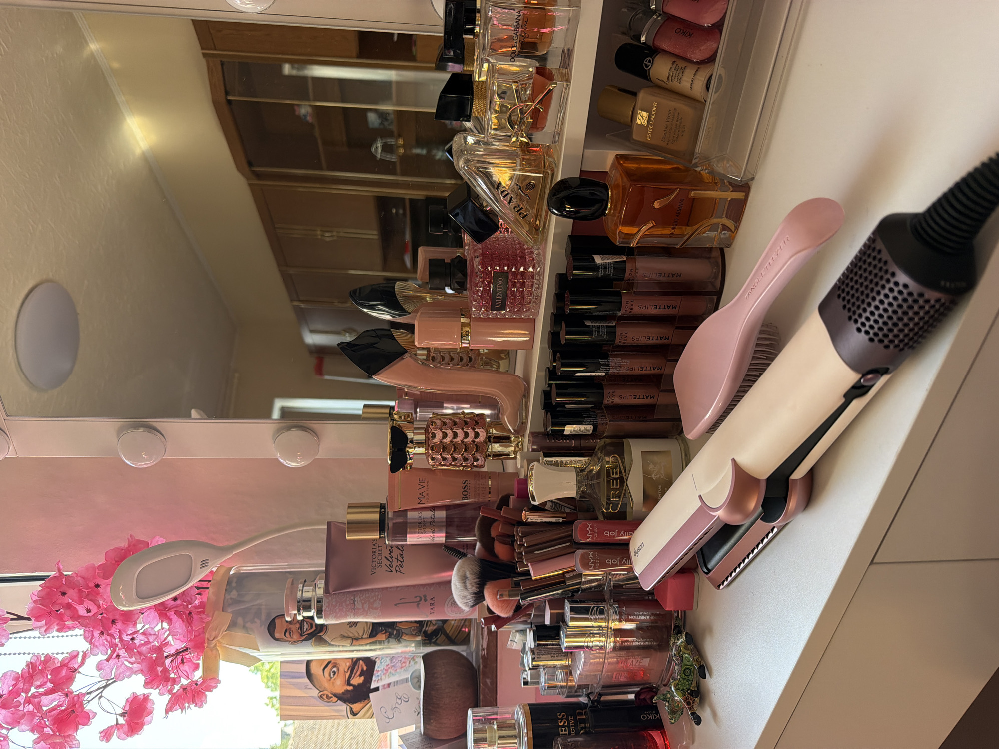 The image depicts a well-lit vanity area filled with makeup and beauty products. In the foreground, a pink and white Dyson straightening iron rests on a white surface, with a pink hairbrush lying next to it. Behind the straightening iron, a variety of makeup items are arranged, including numerous lip glosses, perfumes, and brushes. Notably, several perfume bottles are visible, including brands like "Prada," "Yves Saint Laurent," and "Dolce & Gabbana." A framed picture of two men is propped up on the left side, next to a vase of pink artificial flowers. In the background, a large mirror reflects a room with wooden cabinets. The overall impression is one of a personal grooming space, likely in a home. There is no visual information in the image to suggest it is located in Schwelm, Germany.