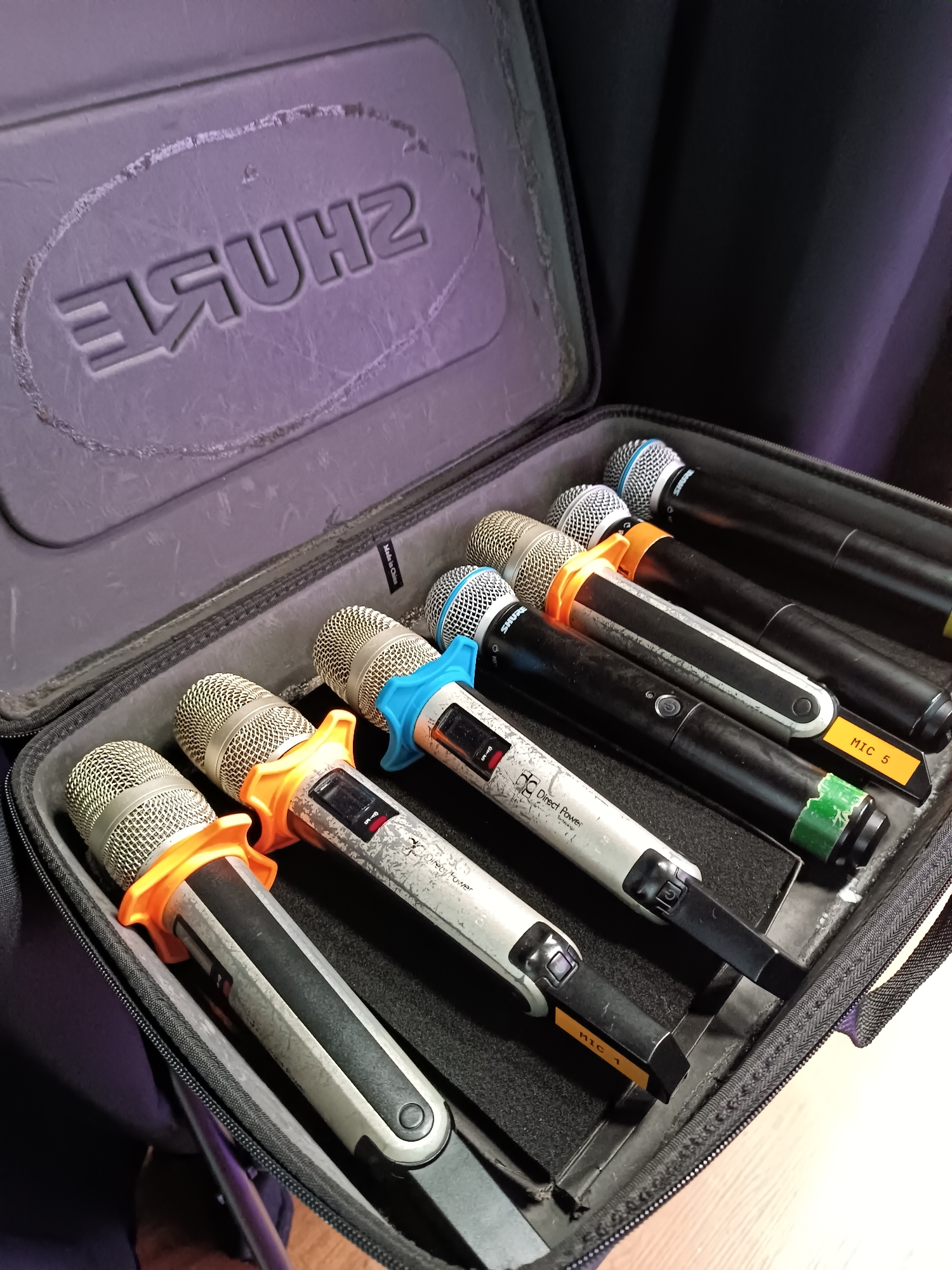 A dark gray Shure brand carrying case is open, revealing multiple wireless microphones. The case has a textured interior and the Shure logo embossed on the lid. There are at least eight microphones visible, mostly black with silver mesh grilles. Some microphones have colored ring accessories around the grilles, including orange and blue. Several microphones have small labels on them, one clearly reading "MIC 5" on an orange sticker. The microphones appear to be in a storage configuration within the case. The image is taken from a slightly elevated angle, looking down into the case. The background is dark and out of focus. The overall scene suggests a professional audio equipment setup. The location is identified as Bykovo, Russia.
