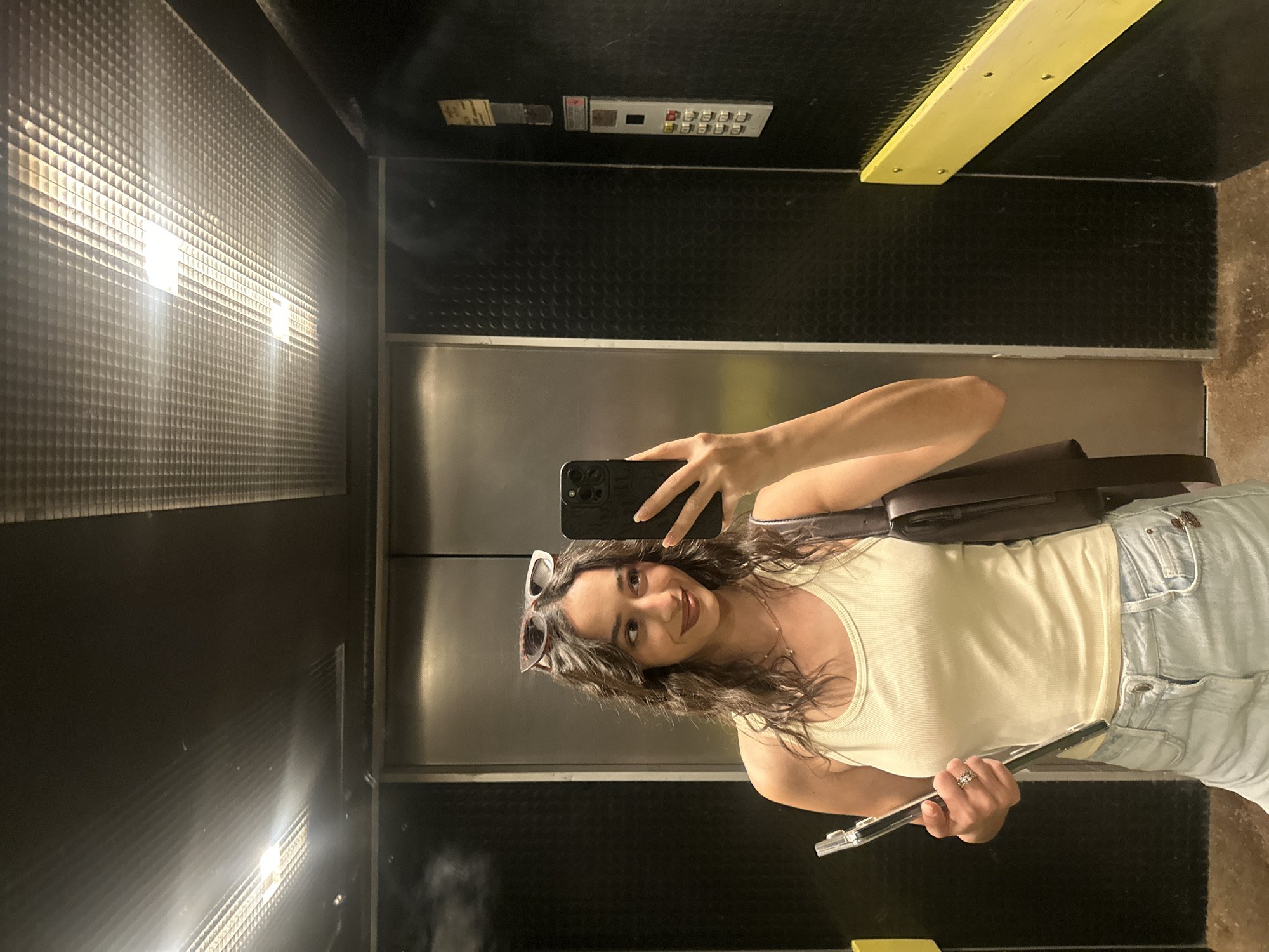 A woman is taking a selfie in an elevator. The elevator interior has black textured walls and ceiling, a stainless steel door, and a yellow handrail. An elevator control panel is visible on the wall. The woman is wearing a light-colored tank top and light blue jeans. She is holding a tablet or slim laptop in her left hand and a smartphone in her right hand to take the picture. A pair of sunglasses rests on top of her head. The location is not discernible from the image.