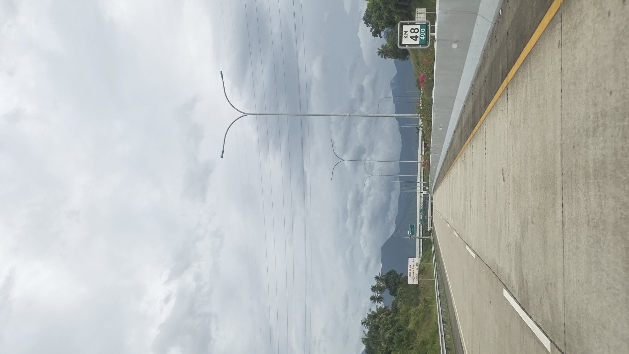 The image depicts a segment of highway in Lampanahtunong, Indonesia. The road surface is concrete, marked with white lines and a yellow line along the edge. Overhead are multiple light poles with dual curved arms, and a network of power lines extends across the sky. In the background, a mountain range is visible, partially obscured by clouds. Lush vegetation including palm trees and other greenery is present on the sides of the road. Road signs are present, including one indicating "KM 48 400". The sky is overcast.