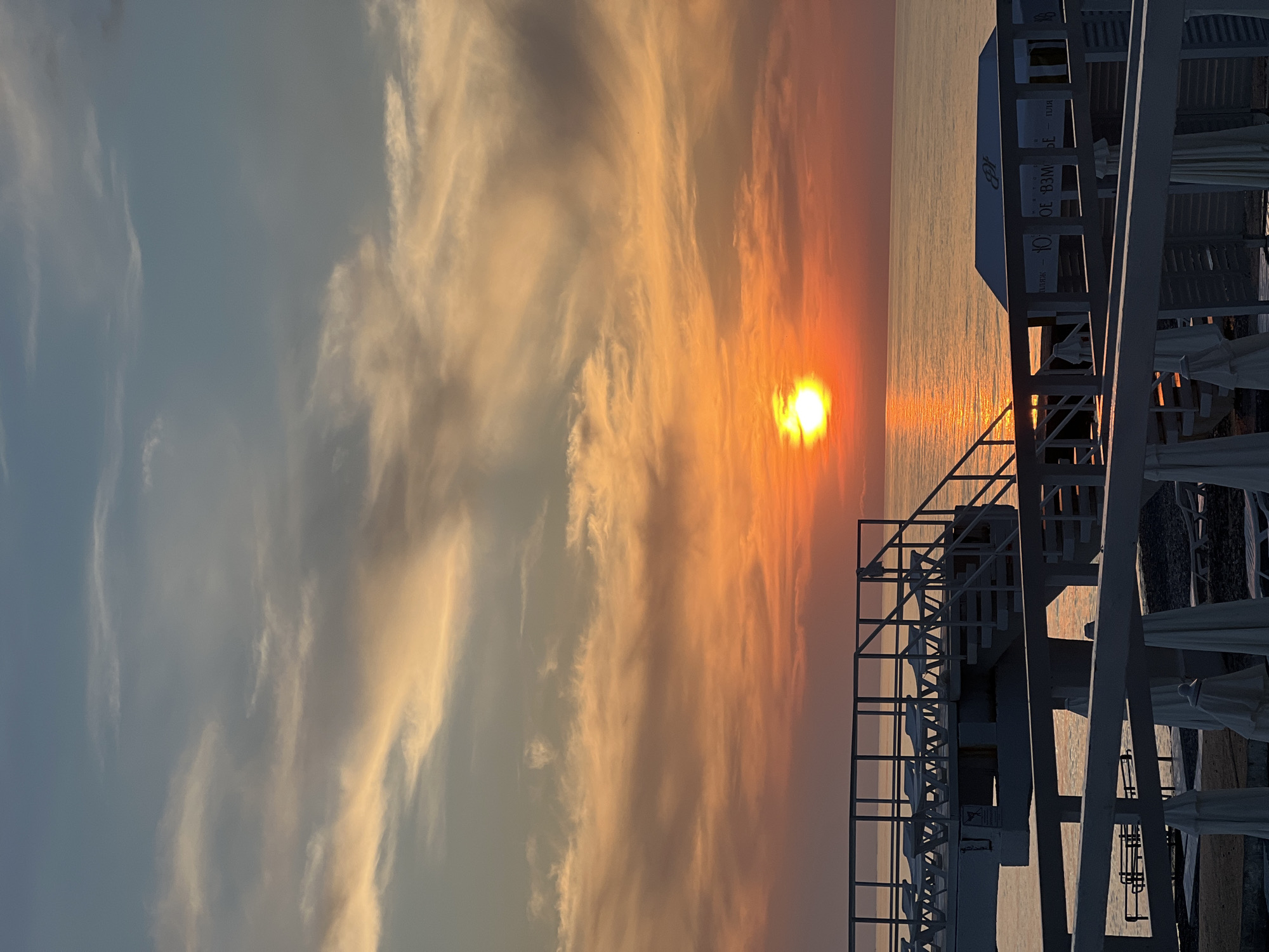 The image depicts a sunset over the Black Sea, viewed from the beach area of the Yuzhnoe Vzmorie Sanatorium in Adler, Sochi, Russia. A bright, glowing yellow-orange sun is positioned low on the horizon, slightly left of the image's vertical center, casting a shimmering path of light across the calm sea. The sky transitions from an intense orange and red near the horizon to lighter orange, pink, and grey-blue towards the top, with scattered cirrus and altocumulus clouds illuminated by the sun's low angle.
In the mid-ground, a multi-level white pier structure extends from the left side into the water, featuring prominent white railings and several sets of stairs leading up and down. The upper deck of this pier holds multiple unoccupied white lounge chairs arranged in rows. A thick white horizontal railing occupies the immediate foreground, partially obscuring the view of the beach level below. This lower level reveals a pebble beach with several folded white beach umbrellas and additional unoccupied lounge chairs. To the right, a white building, likely a beach cabana or pavilion, is visible with a peaked roof. Its side displays mirrored text that reads "ЮЖНОЕ ВЗМОРЬЕ" (Southern Seashore) and "ПЛЯЖ" (Beach), confirming the location. No individuals are present in the scene.