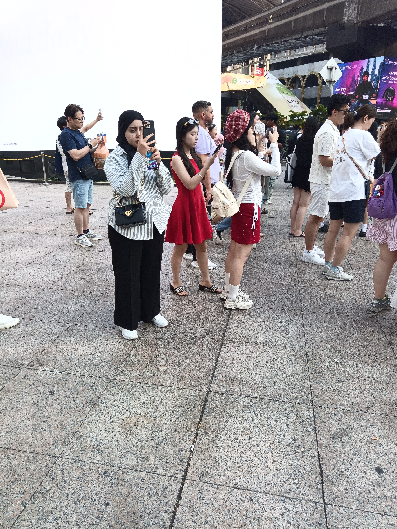 A diverse group of people are standing on a paved walkway in Kuala Lumpur, Malaysia, near a large white screen and an elevated train line. Many are holding up their mobile phones, presumably to take photos or record videos, suggesting a public event or attraction.
In the foreground, a woman wearing a black hijab and a striped long-sleeved shirt is holding her phone up to take a picture. She has a black purse with a gold clasp. Beside her, a woman in a red dress is also holding a phone.
Further back, a man in a blue t-shirt and glasses is holding his phone, and a man in a white t-shirt and shorts is looking towards the screen. To the right, other individuals are visible, some wearing casual clothing like t-shirts and shorts, and one person is carrying a purple backpack. The environment appears to be an urban outdoor setting with overhead structures and advertising billboards in the background. The ground is made of large granite tiles.