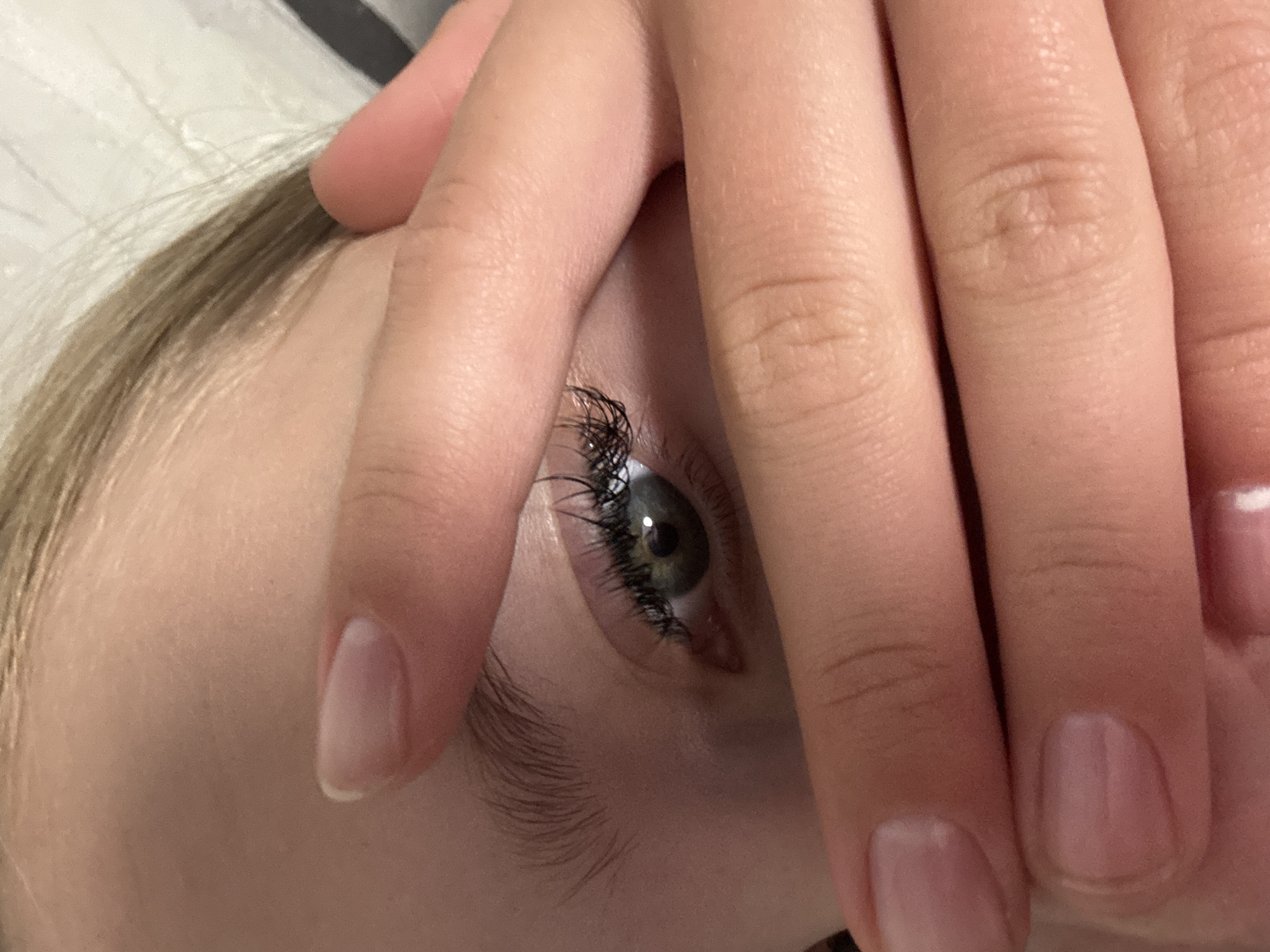 The image is a close-up, angled shot of a person's eye, partially obscured by their fingers. The eye is green with dark eyelashes. The skin is fair, and blonde hair is visible in the upper right corner of the frame. The fingers are also fair-skinned and appear to be resting against the person's face. The background is out of focus and appears to be a textured surface, possibly a bedsheet. There is no discernible location in the city of Chaikovsky, Russia, or any other specific environmental context provided by the image.
