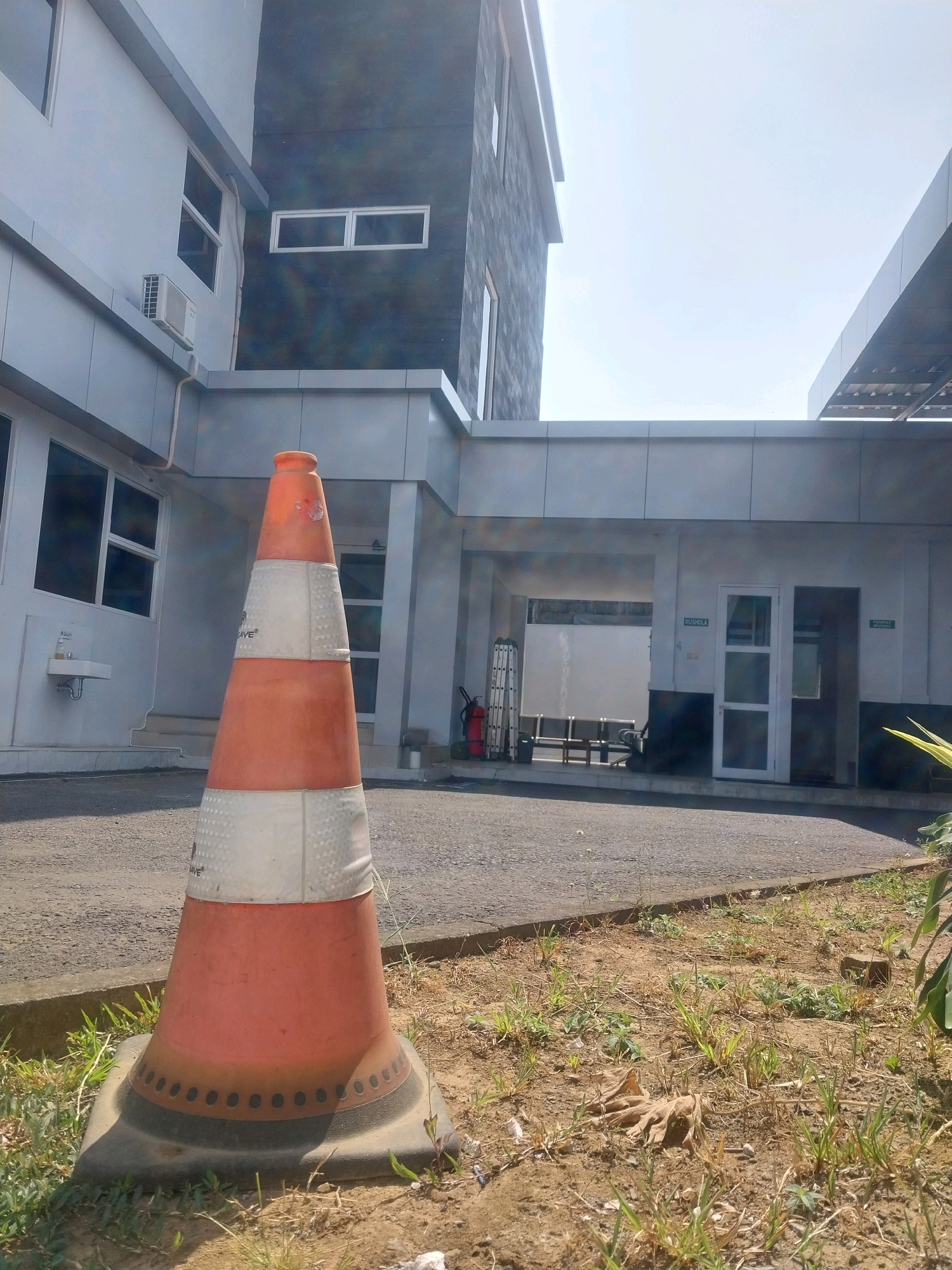 A low-angle, eye-level outdoor shot captures a brightly lit, modern building complex with a prominent orange and white traffic cone in the foreground. The building, likely located in Sleman, Indonesia, features a mix of grey and dark grey facades with white window frames. To the left, a white section of the building has windows and an outdoor air conditioning unit. The central section of the building is darker and taller, with horizontally aligned windows. A covered walkway connects the buildings.
The traffic cone, a focal point of the image, is positioned on a paved area bordered by dry grass and dirt. It is conical in shape, with reflective white bands alternating with orange sections. The base of the cone is wide and black.
In the background, through an open doorway, a white partition wall is visible, along with several rows of benches. A red fire extinguisher and a collapsed silver ladder are leaning against the wall near the entrance. Green signs are affixed to the building; one reads "MUSHOLA," and another, to the right, says "TEMPAT WUDHU." The sky is clear and bright. The overall impression is one of an institutional or commercial setting on a sunny day.
