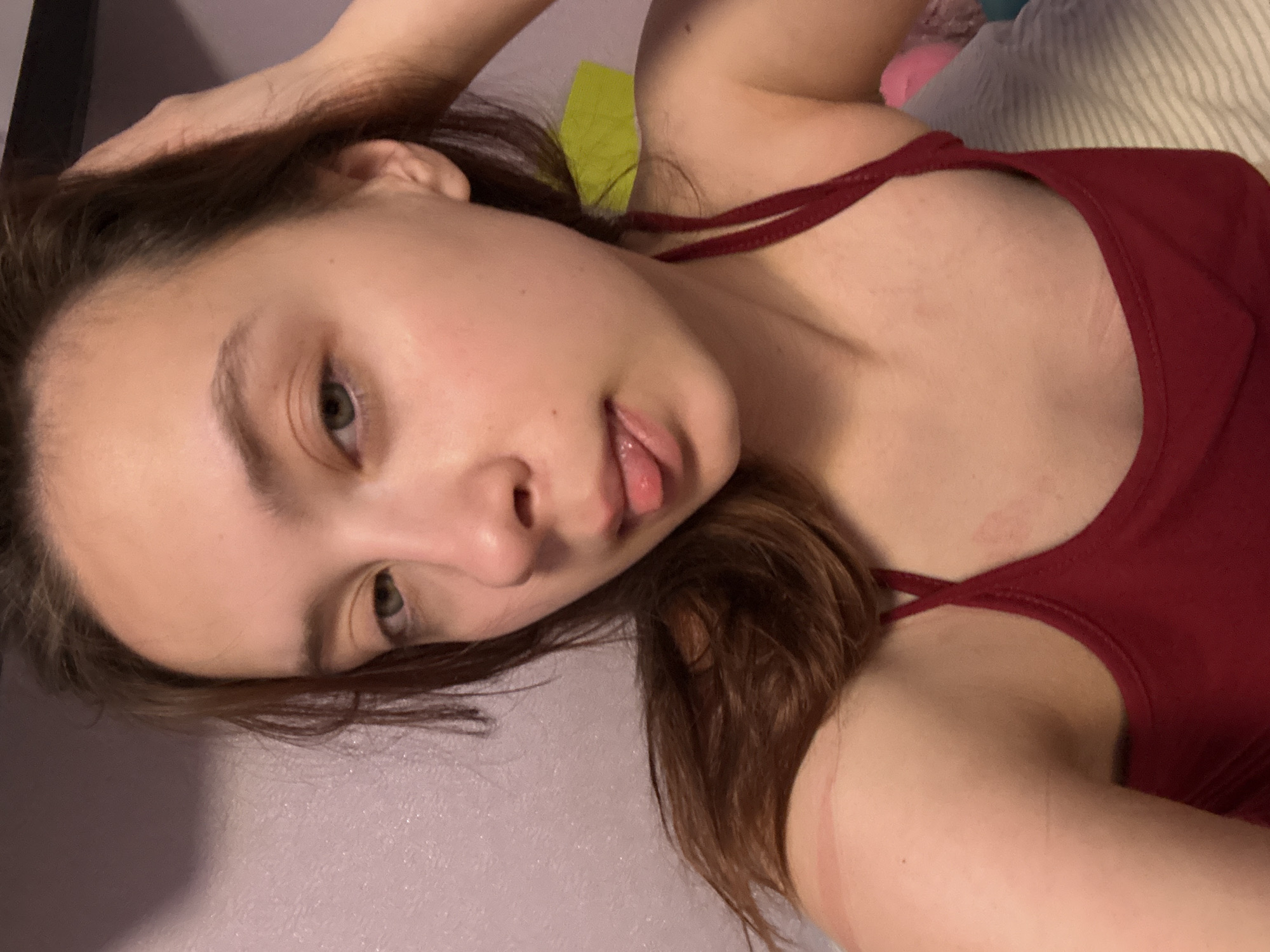 A young woman with light skin and brown hair lies on her side, facing the camera. She is wearing a red tank top with thin straps. Her tongue is slightly extended. The background is a wall with a light lavender or pink hue. Other objects in the background include a striped pillow, a pink stuffed animal, and a lime green object. The location is Przykamenskyj, Russia, though there are no indicators of this location within the image.
