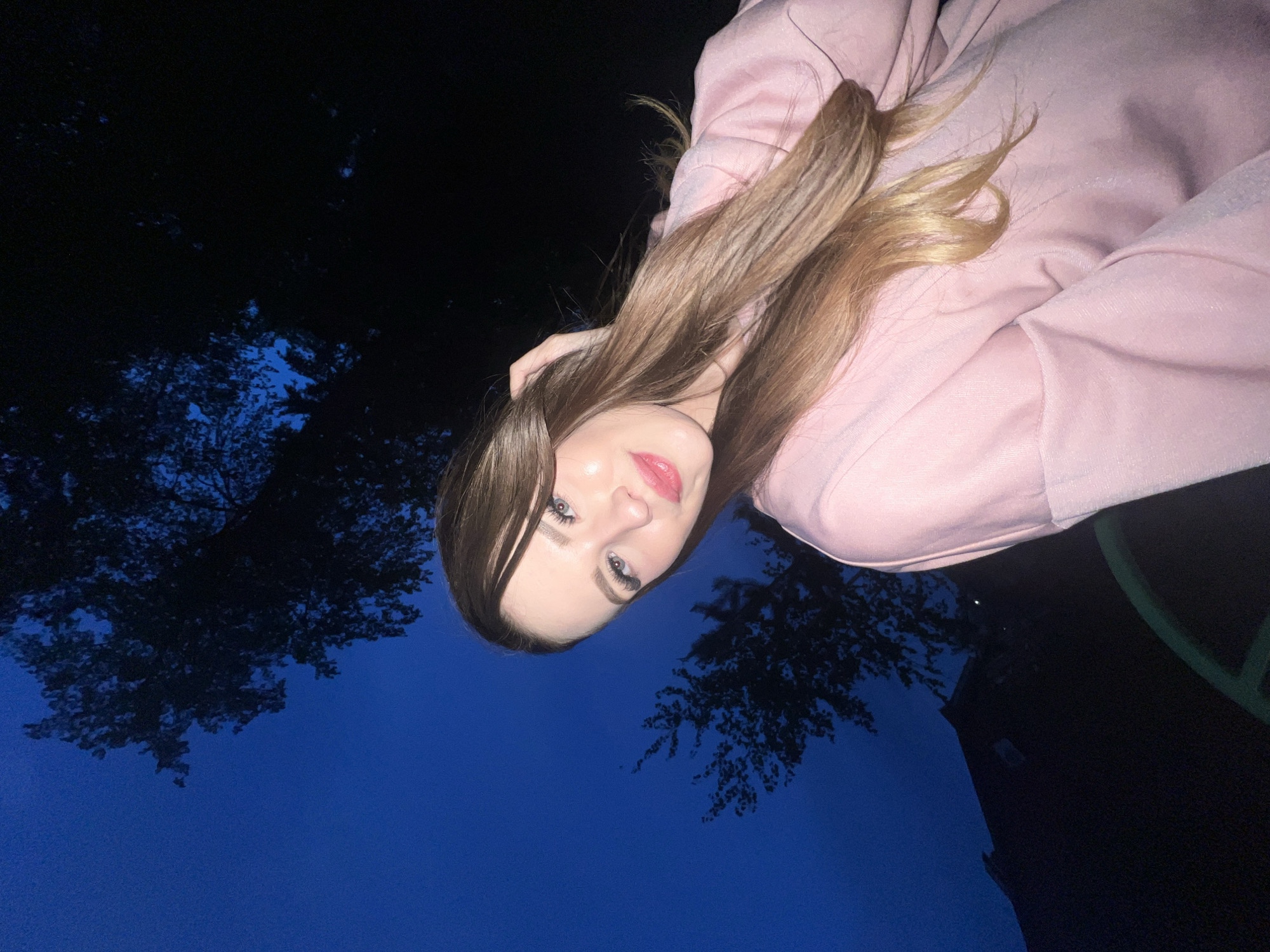 A woman with long, brown and blonde hair, wearing a light pink sweatshirt is the main subject. She is positioned in the center of the frame, angled towards the camera. Behind her is a dark blue sky, interspersed with tree branches. A building is visible in the right part of the image. The image appears to be taken outdoors at night. The location information is not discernible from the visual data of the image.