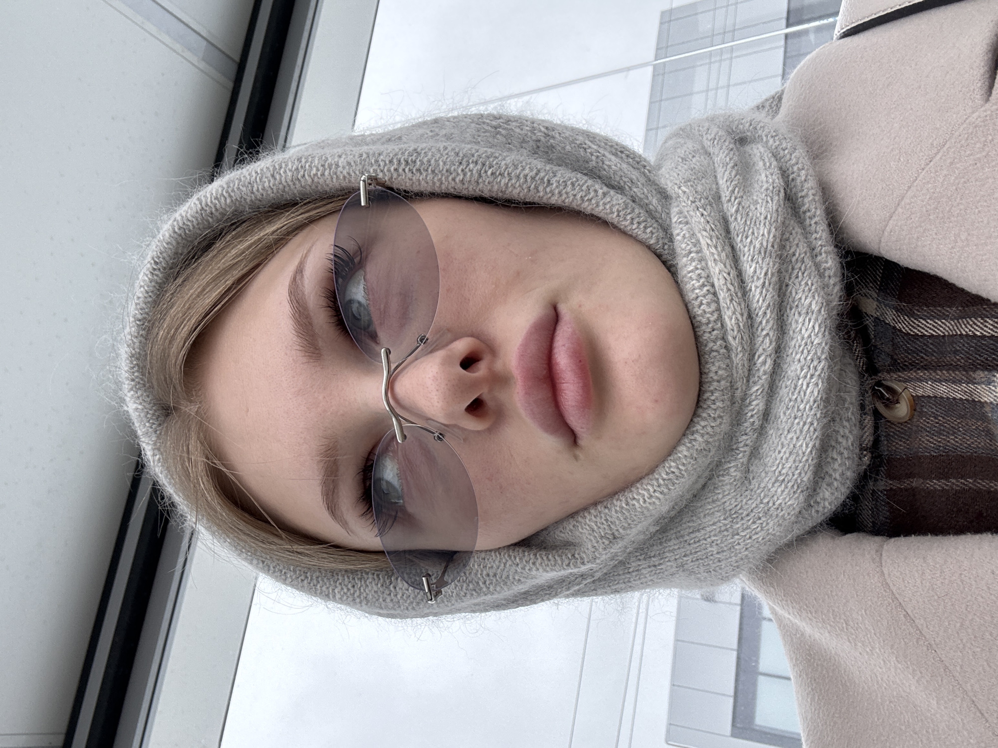 The image captures a close-up portrait of a young person, likely female, facing forward in a slightly tilted vertical orientation. The subject has fair skin, light-colored hair visible around the forehead, and light blue-grey eyes. She wears rimless, light-tinted oval sunglasses that cover her eyes, revealing long eyelashes through the lenses.
She is dressed in warm clothing, featuring a soft, thick, light-grey knitted hood or scarf that frames her face and neck. Over this, she wears a beige or cream-colored coat, and a brown and cream plaid garment is visible underneath the coat on her right side.
The background, seen through a large window, depicts an overcast sky and distant architectural structures, including window frames and building outlines, suggesting an urban environment. The light outside indicates daytime, and the warm clothing implies cooler weather, consistent with the Perm, Russia context. The subject maintains a neutral, relaxed expression while looking towards the camera. There is no visible text in the image.