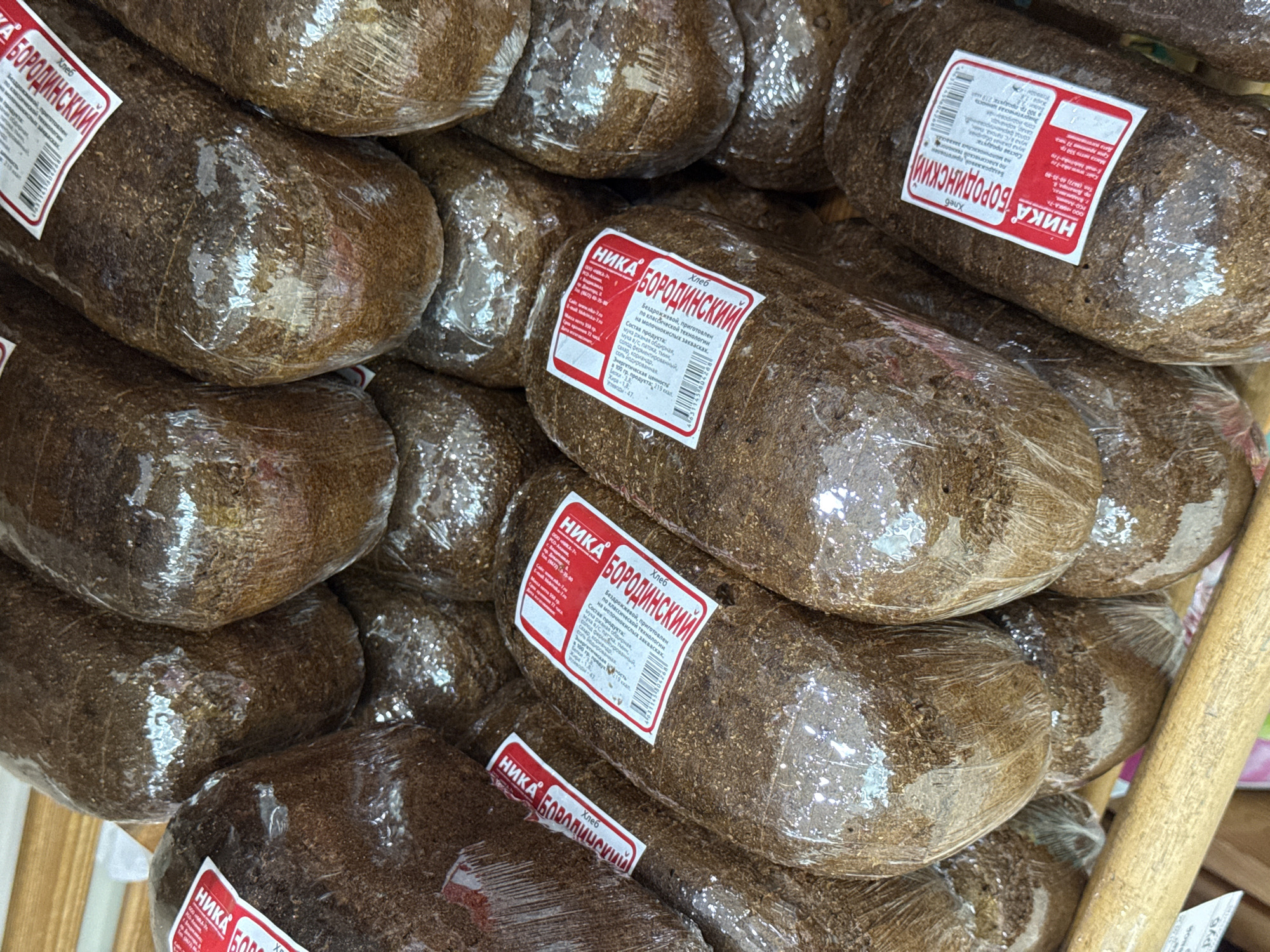 This is a close-up, overhead shot of a stack of dark, oval-shaped loaves of Borodinsky bread, a Russian rye bread. The loaves are individually wrapped in clear plastic. Each loaf has a red and white label with Cyrillic text. The labels indicate the brand "НИКА" (Nika) and the bread type "БОРОДИНСКИЙ" (Borodinsky). Some labels also provide information about the ingredients, nutritional value, and manufacturer. The bread appears to be on display, possibly in a bakery or market stall, with some wooden elements visible in the background. The lighting suggests an indoor setting, possibly during the day. The focus is entirely on the bread, with no people or other objects clearly visible. The overall impression is one of abundance and a focus on a traditional food item.