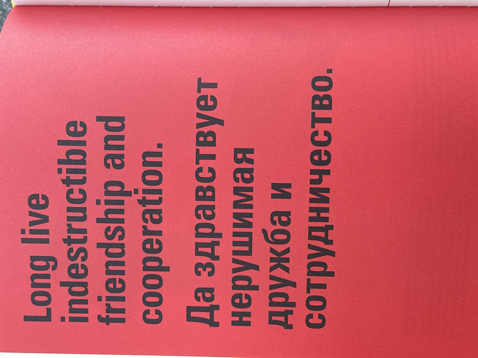 The image presents a single, brightly colored red page from a publication, located at an unspecified indoor or outdoor setting within the city of Tel-Aviv, Israel. The page features prominent black, sans-serif text in two languages. The upper portion displays the English phrase: "Long live indestructible friendship and cooperation." The lower portion contains the identical phrase in Russian Cyrillic script: "Да здравствует нерушимая дружба и сотрудничество." The page exhibits a slight curvature along its right edge, revealing the spine of a bound document or book, with a thin, light pink strip of an adjacent page visible along the binding line. No individuals, actions, or specific environmental features beyond the page itself are discernible.