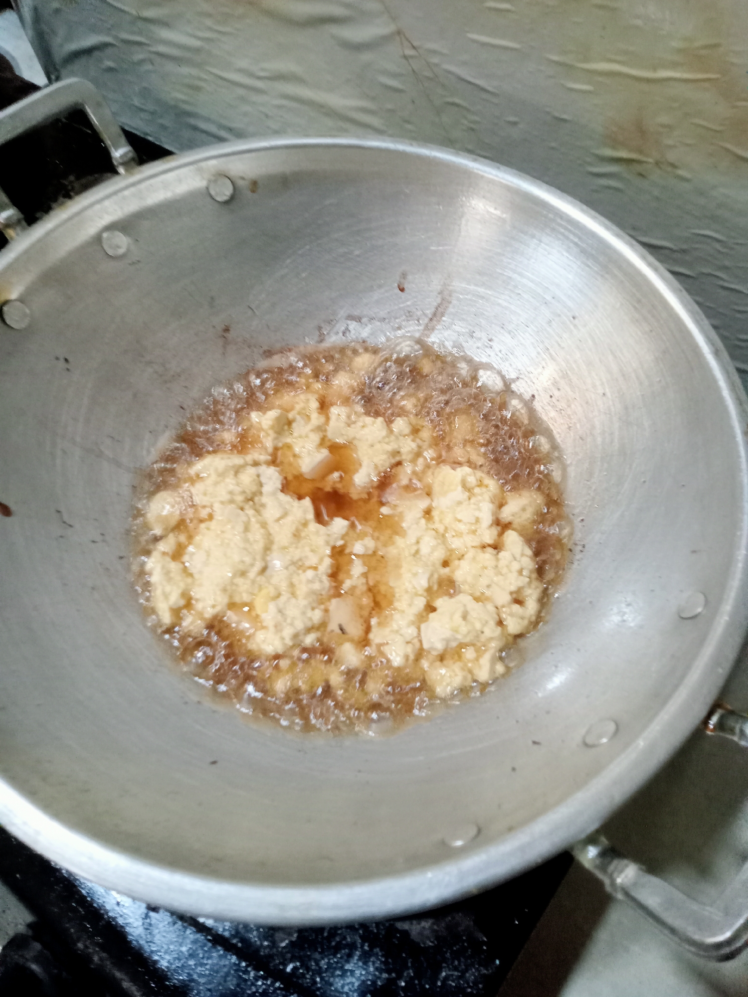 In Simo, Indonesia, a close-up aerial shot captures a meal being prepared in a metal wok. The wok, filled with bubbling hot oil, contains scrambled eggs mixed with small pieces of what appear to be vegetables or meat. The eggs are partially cooked, with some areas still pale yellow and others taking on a golden-brown hue from the oil. The oil itself is actively frying, creating a foamy texture around the food. The wok is placed on a dark, stovetop surface. In the background, a light-colored, textured surface is visible, possibly a wall or a cloth. The overall scene depicts a home kitchen environment where food is being cooked.