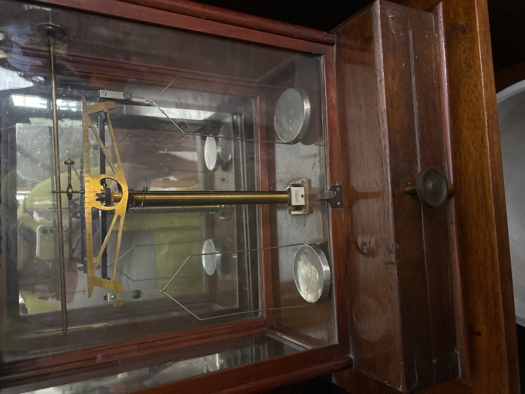 The image shows an antique balance scale enclosed in a glass and wooden display case, which is placed on a wooden surface. The balance scale is made of brass and has two circular pans suspended by wires. A central beam with markings is visible above the pans, along with a pointer. The display case has wooden framing and glass panels, with a drawer at the bottom. The overall scene suggests a static display of historical equipment, likely in an indoor setting such as a museum or an antique shop. The lighting is somewhat dim, casting shadows and highlighting the reflective surfaces of the scale and the glass.
