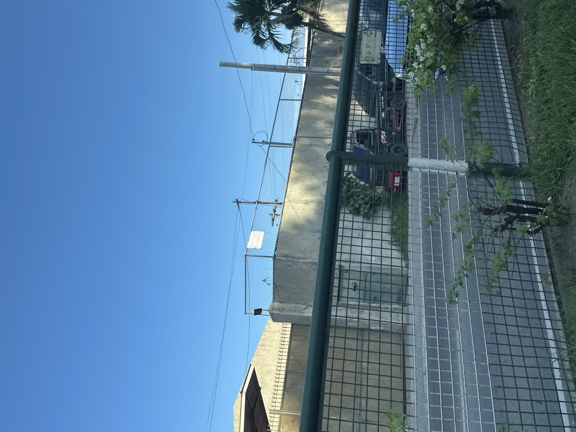 This image depicts an outdoor scene in Davao City, Philippines, under a clear, bright blue sky. The foreground is dominated by a dark green metal mesh fence with a grid pattern. Immediately in front of and partially through the fence, there is green grass and small shrubs with light green leaves and clusters of white flowers. A white vertical post, part of the fence structure, is visible in the lower center-right.
Behind the fence, a paved asphalt road or path extends horizontally, marked by a distinct solid white line parallel to the fence. Further back, a tall, light gray concrete wall forms a prominent barrier, topped with a chain-link fence that runs the length of the visible area.
Above the concrete wall and chain-link fence, various elements are visible. On the left, a building with a light-colored, possibly sloped roof and a matching wall is partially seen, along with a dark, ornate metal gate featuring spiked top elements. Towards the center-left, a white rectangular sign with black text is affixed to the chain-link fence, displaying "DIRECT SALE" and the phone number "0925834605". A small, dark, rectangular device is mounted on a short pole just below this sign, rising from the concrete wall. Multiple electrical wires traverse the upper part of the scene horizontally.
On the right side, a tall, slender, light gray utility pole stands vertically, and a green palm tree is partially visible on the far right. Through the fence and above the road, two vehicles are partially discernable. One appears to be a utility vehicle or truck, colored red and black. To its right, a blue vehicle is parked. A white sign bearing the text "D.Y.C. LIFE IS HERE" is attached to the fence near the blue vehicle. The overall lighting suggests daytime, with shadows indicating the sun is at an angle. There are no people visible in the scene.