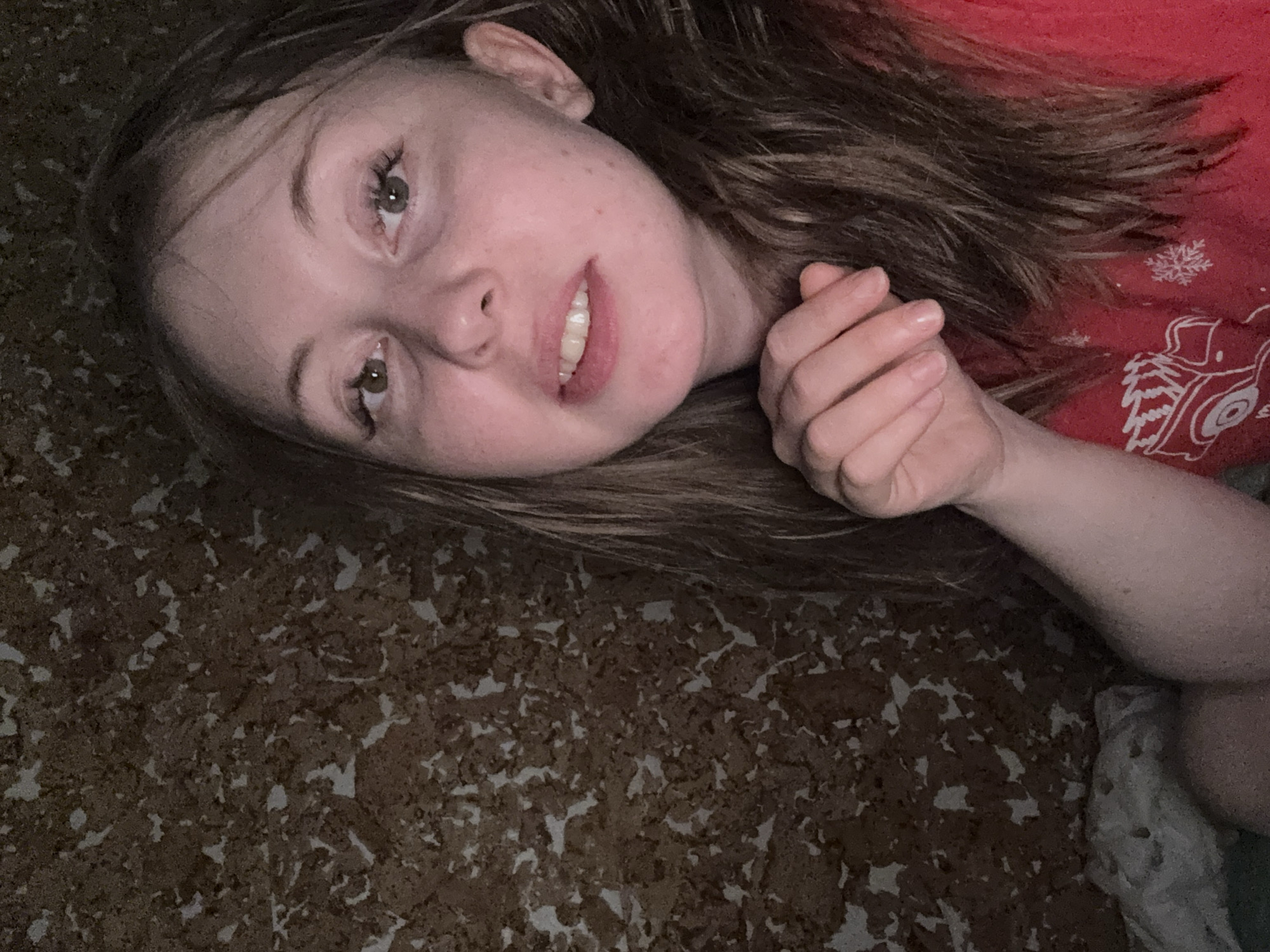 A young fair-skinned woman with brown hair is lying down. Her head is tilted towards the upper left corner. She has light brown eyes and is slightly smiling. Her right hand is clenched near her cheek. She is wearing a red shirt with white graphics. The surface she is lying on appears to be a mottled brown and white material. The location is Unknown, Unknown.