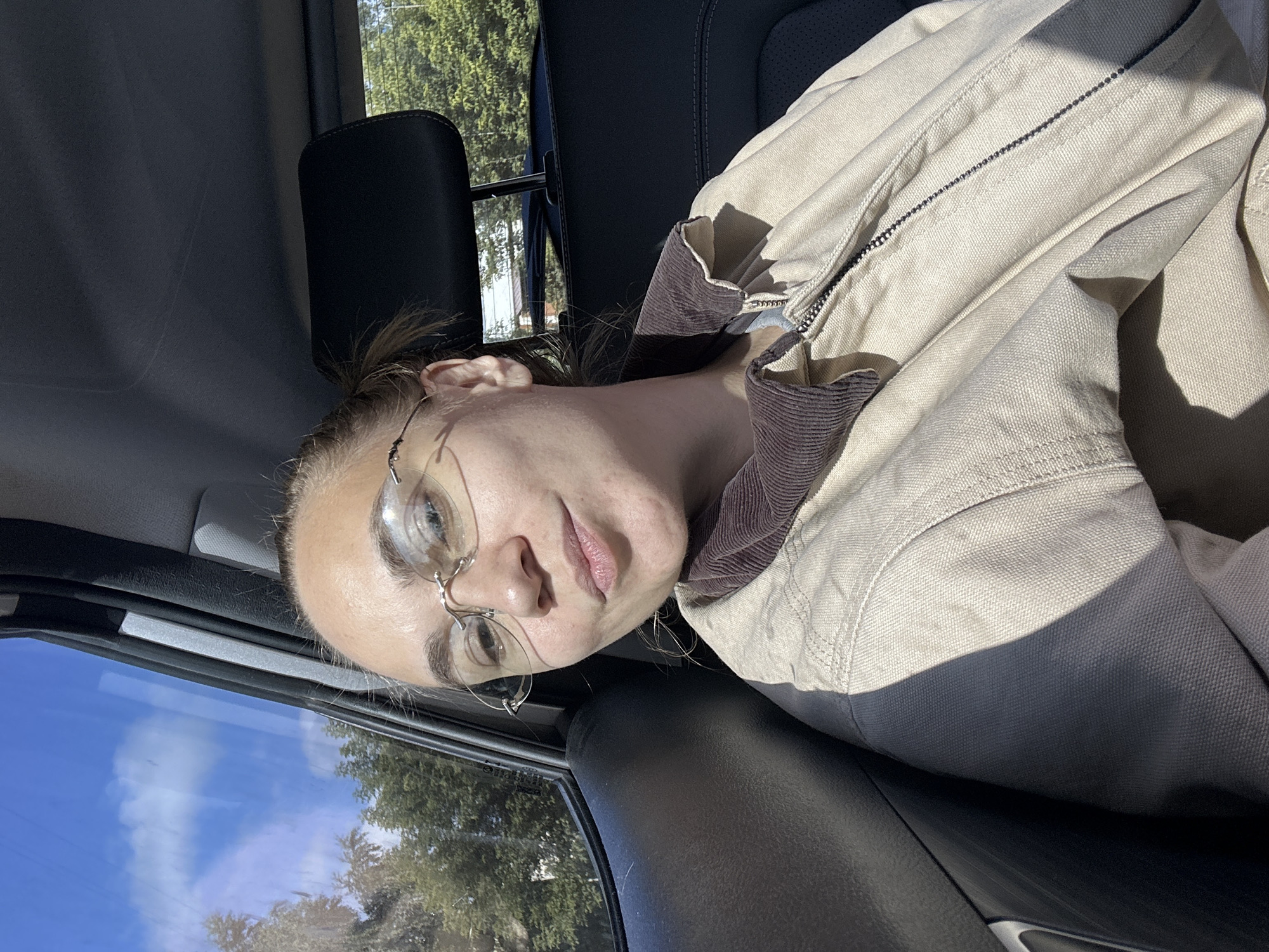 A medium shot photograph captures a woman with light skin and brown hair pulled back in a bun, wearing round eyeglasses and a tan jacket with a dark brown corduroy collar. She is seated in the passenger seat of a vehicle, looking towards the camera with a slight smile. The window beside her reveals a bright blue sky with scattered clouds and the tops of green trees, indicating an outdoor environment. The interior of the car is visible, including a dark gray headrest and seat. Sunlight streams in, casting shadows across the woman and the car's interior. The scene appears to be in a suburban or natural setting, possibly outside the city of Chaikovsky, Russia, given the context.