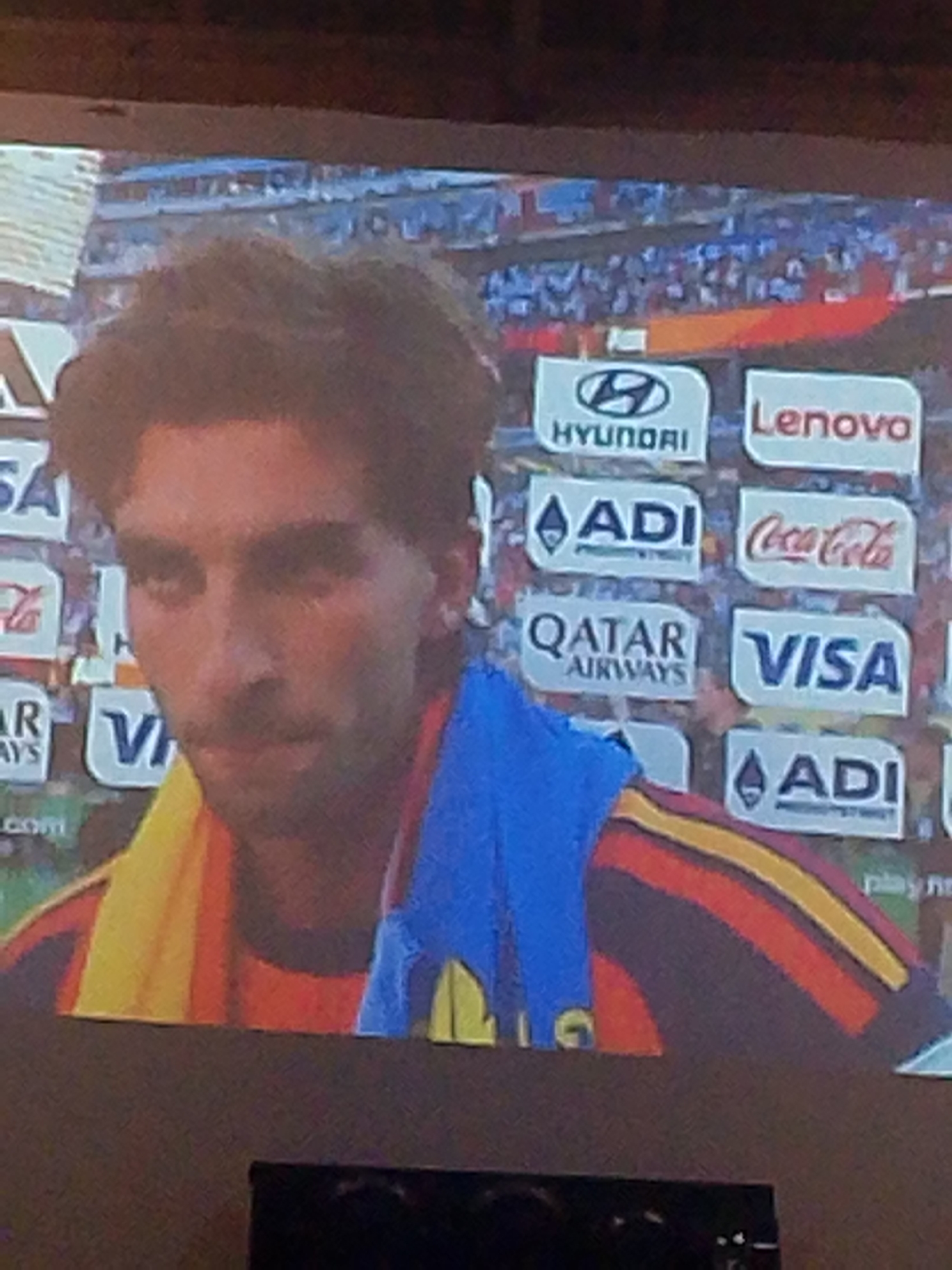 A medium shot captures a close-up of a man's face, with the top half of his body visible. He has dark, wavy hair and a thin mustache and stubble. He is wearing a blue towel draped over his shoulders and a jersey with red, yellow, and black stripes. Behind him, a wall of advertisements is visible, featuring logos for Hyundai, Lenovo, ADI, Coca-Cola, Qatar Airways, and Visa. The background appears to be a stadium with a crowd, suggesting a sporting event. The image is blurry and appears to be taken from a distance or on a screen, with a dark border at the bottom suggesting it's a photograph of a television. No specific location in Meunasahmee, Indonesia, can be identified from the provided image.