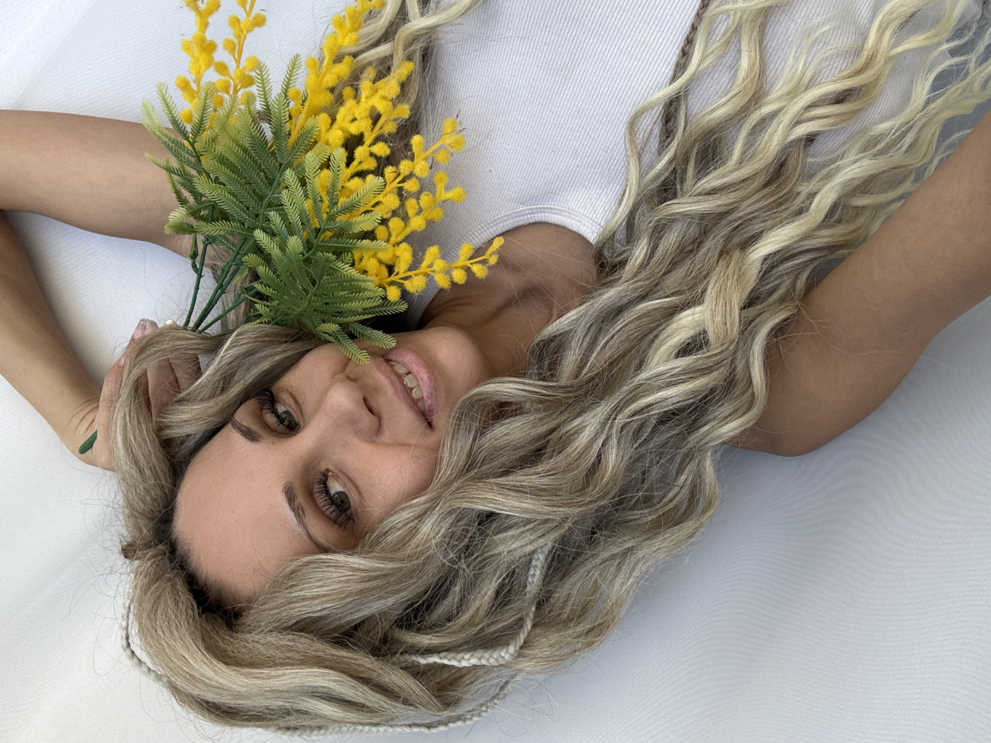 A woman lies on a white surface, her head tilted toward the camera. Her blonde hair is wavy and scattered around her head, and she is wearing a white tank top. She is holding a bouquet of yellow mimosa flowers in her left hand near her head. The background is a plain white surface. The city of Rusanovo, Russia is not discernible from this image.