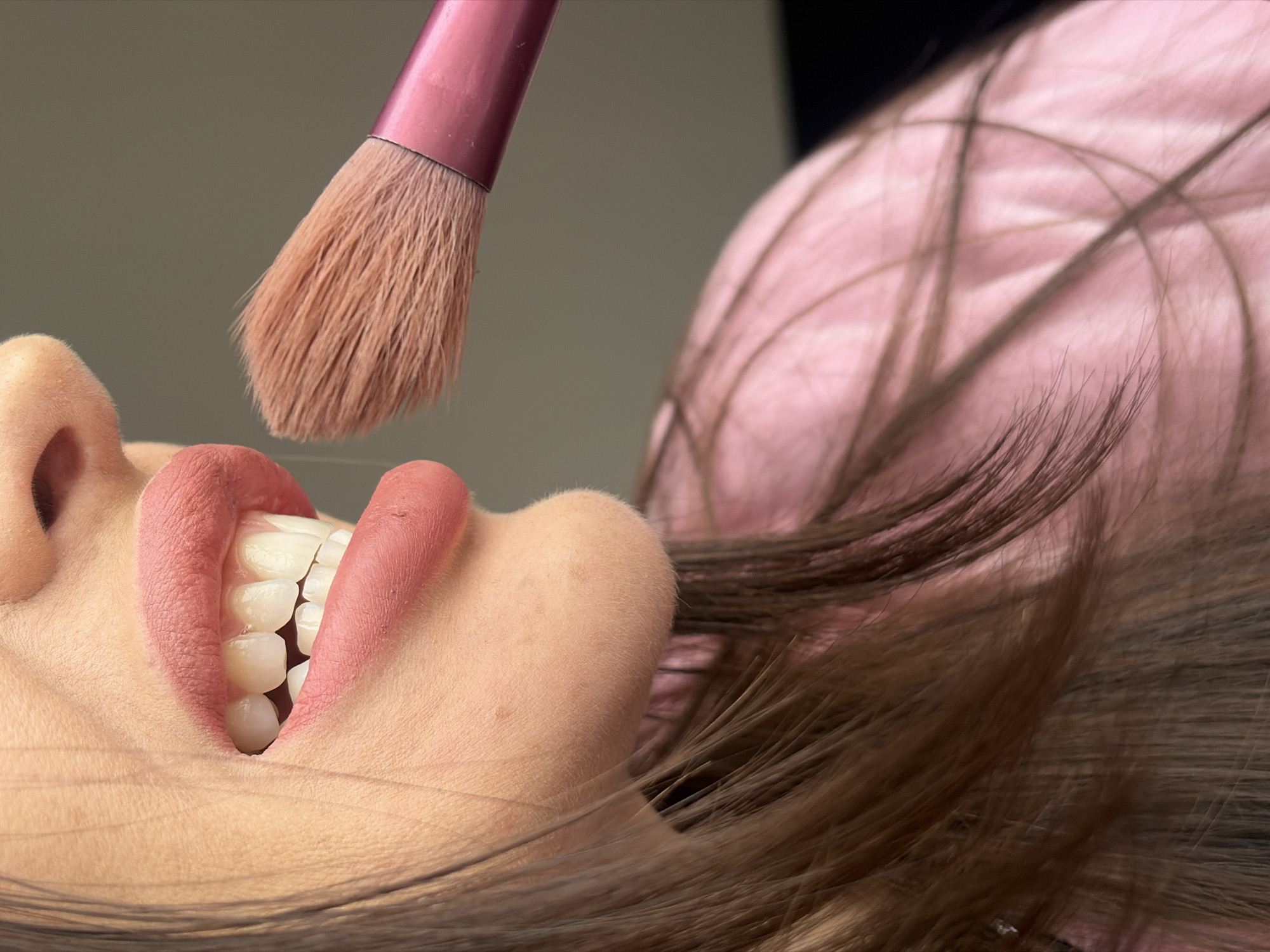 The image depicts a close-up, angled view of a person's lower face, set in Saint Petersburg, Russia. The subject's mouth is slightly open, showcasing healthy, white teeth. The lips are covered in a matte, medium-pink lipstick. The skin around the chin and mouth exhibits a smooth texture, with a few faint blemishes visible on the chin. Brown hair frames the left side of the face and extends towards the lower right, partially covering a pink garment. A makeup brush, featuring light brown bristles and a vibrant pink handle, is positioned to the right of the mouth, indicating an action related to makeup application. The background consists of an out-of-focus, uniform light grey-beige area in the upper right and blurred pink fabric below, consistent with an indoor setting.