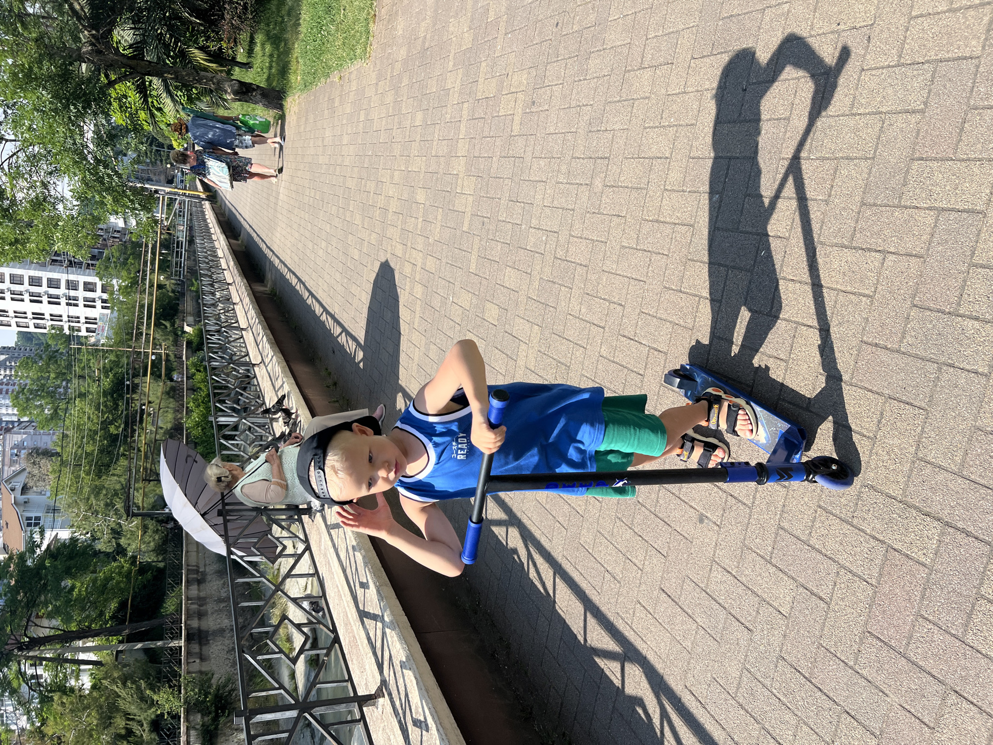 The image depicts an outdoor scene on a sunny day along a paved pathway in Sochi, Russia.
In the foreground, a young blonde boy, estimated to be between 4 and 7 years old, is riding a blue kick scooter. He wears a black baseball cap backward, a blue sleeveless shirt with "READY" printed in white, green shorts, and open-toed sandals. His left hand grips the scooter's handlebar, while his right hand is raised to his ear or adjusting his cap. He looks forward and slightly to his left. A prominent shadow of the boy and scooter is cast to his right on the light-colored interlocking paver pathway.
To the left of the pathway, a black metal fence with geometric patterns separates the path from a narrow, murky waterway, likely a river or canal. Beyond the waterway, green vegetation and buildings are visible. An elderly woman with light hair, wearing a light-colored vest and trousers, is seated on a low wall or bench next to the fence. She holds a large, two-toned (white and dark) umbrella for shade and appears to be observing or interacting with several pigeons situated on the wall near her. Another person, partially obscured, is visible behind the boy's head near the fence.
Further down the pathway on the right side, two individuals are walking away from the viewer. One person wears a patterned dress and carries a light-colored bag. The other wears a patterned shirt, dark shorts, and a hat, carrying a green bag. The pathway is bordered by a grassy area with trees and bushes on the right. In the background, tall residential or commercial buildings are visible, including a multi-story white building with numerous windows. Overhead wires and cables traverse the upper portion of the scene.