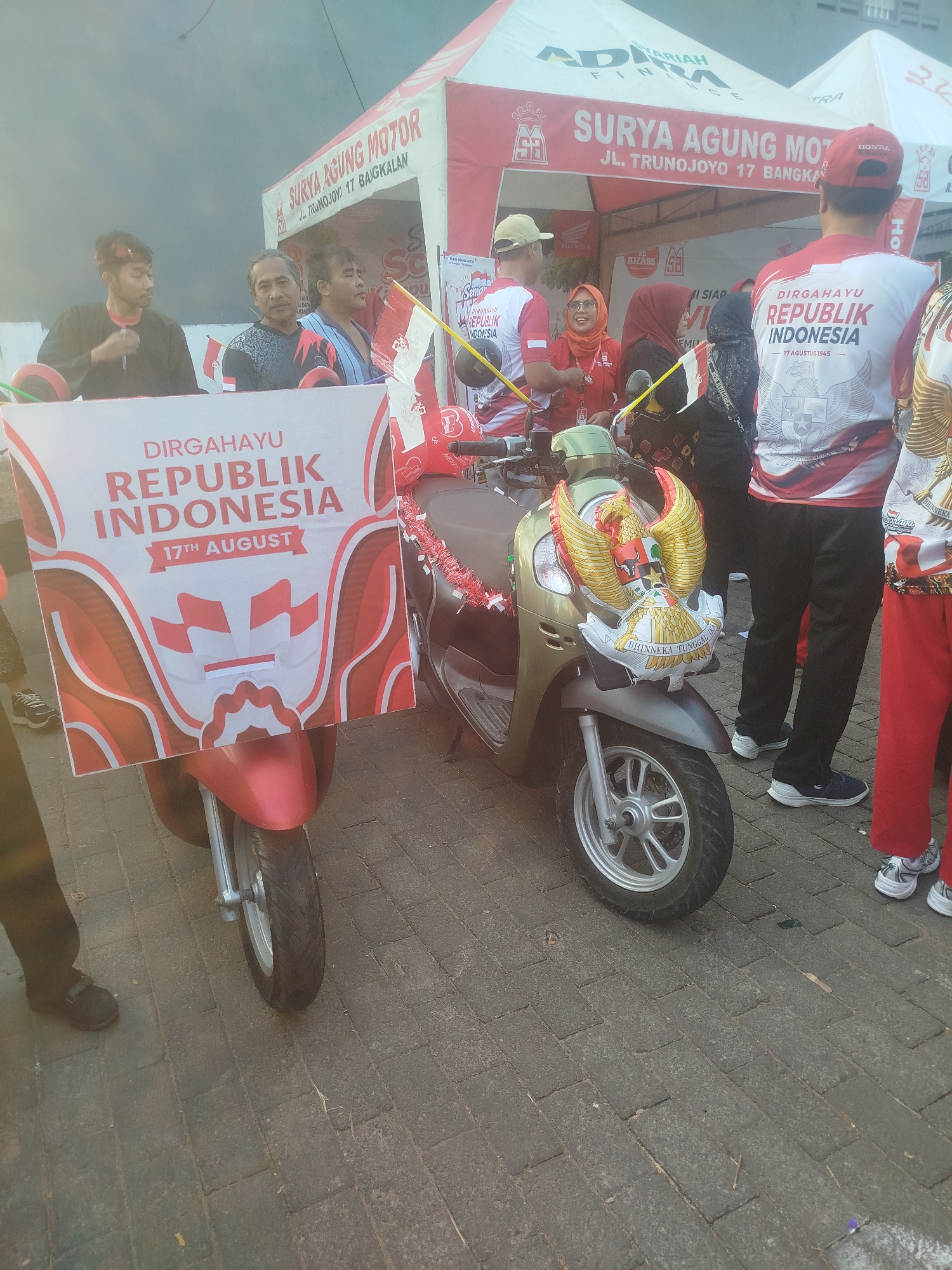 The image captures a scene in Burneh, Indonesia, featuring a street decorated for Indonesian Independence Day. In the foreground, a red scooter is parked. A large banner on the scooter reads "DIRGAHAYU REPUBLIK INDONESIA 17TH AUGUST" with a design of Indonesian flags. A gold and red balloon in the shape of Garuda Pancasila, the national emblem, is attached to the front of the scooter, with "BHINNEKA TUNGGAL IKA" written below it. Behind the scooter, a tent with "SURYA AGUNG MOTOR" and "JL. TRUNOJOYO 17 BANGKALAN" printed on it is visible, suggesting it is a motorcycle dealership. Several people are present, some wearing red and white attire, indicating participation in the celebration. One person in the background is wearing a red cap with a Honda logo. The ground is paved with interlocking bricks. The overall atmosphere suggests a community event or parade celebrating Indonesian independence.
