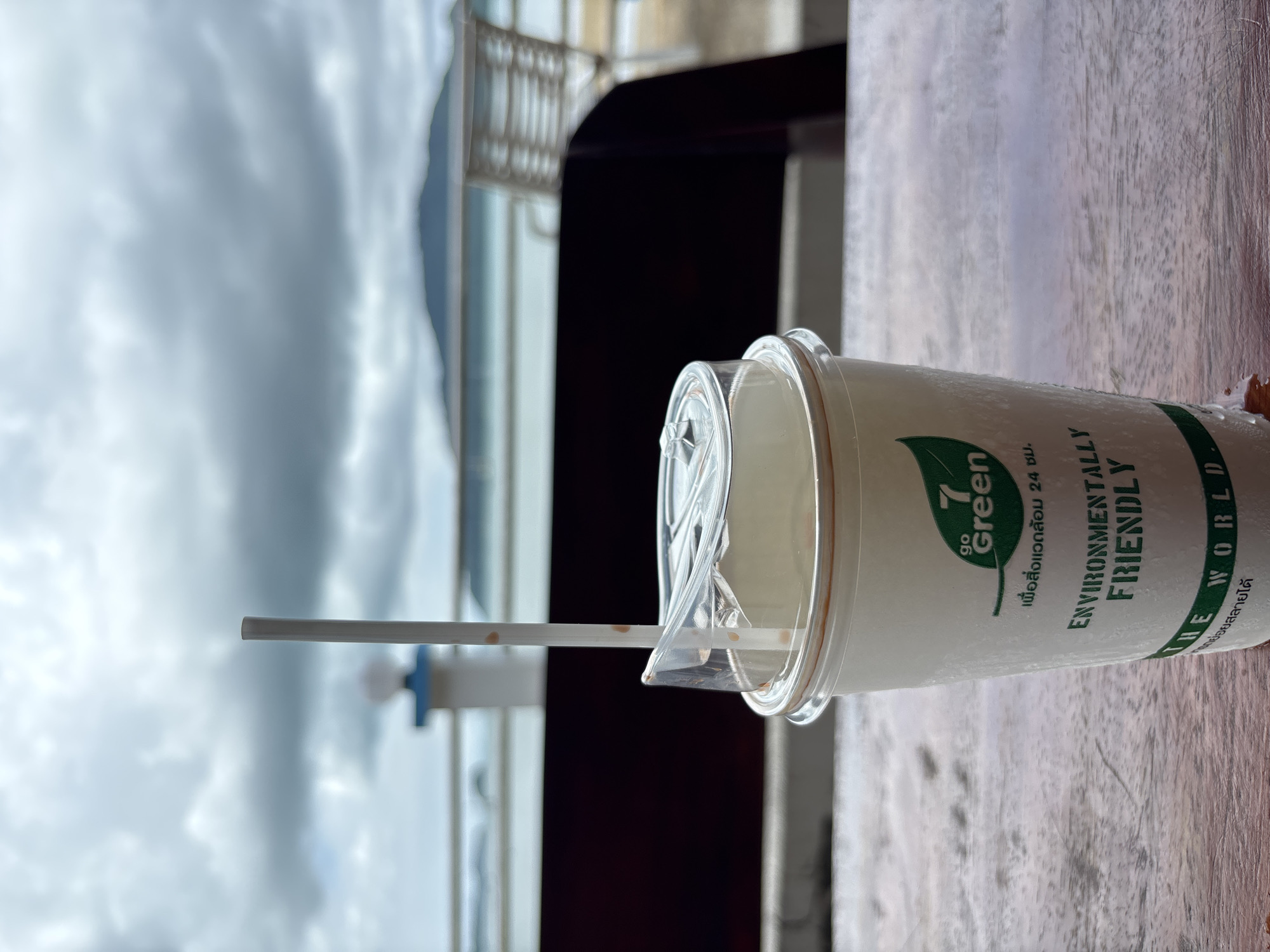 A white paper cup with a clear plastic lid and a paper straw rests on a wooden table. The cup has green text that reads "7 go Green" and "ENVIRONMENTALLY FRIENDLY THE WORLD". The cup is beaded with condensation. In the background, out of focus, is a beach scene with the ocean, cloudy sky, and distant mountains. The location is Rawai, Thailand.