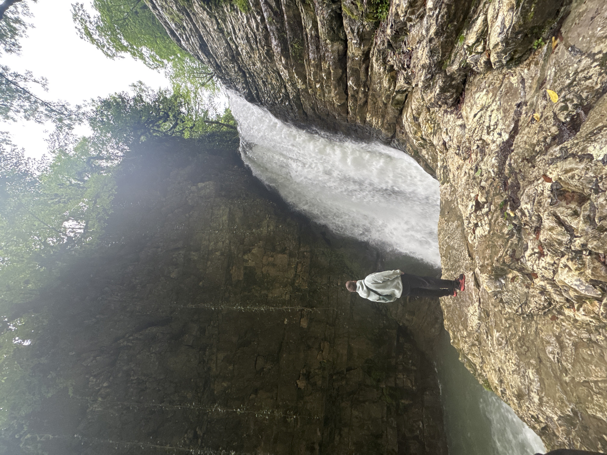 A medium, eye-level shot captures a person positioned on a rocky outcrop in front of a waterfall. The person is wearing a light-colored hooded sweatshirt, dark pants, and red shoes. They are facing the waterfall with their hands in their pockets. The waterfall is white and appears to be forceful. The surrounding environment consists of steep, rocky cliffs covered with greenery, indicative of a natural setting. Based on the location provided, this scene is likely in or near the city of Tsabal, Abkhazia.