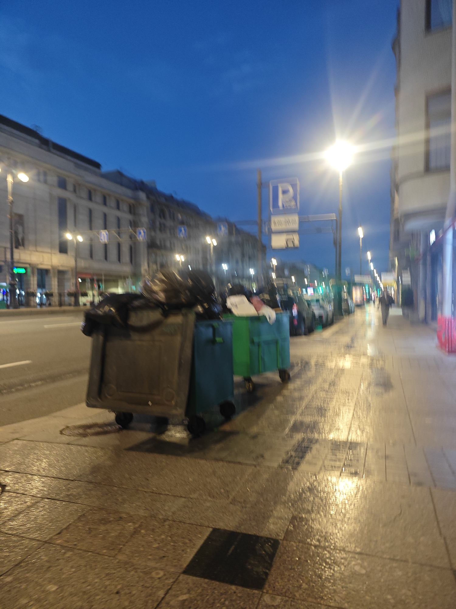 The image captures a street scene at dusk in Saint Petersburg, Russia, likely on a side street or a less busy avenue. The sky is a deep blue, indicating twilight. A street lamp with a bright, star-like glow illuminates the scene. In the foreground, two large, dark green trash bins are overflowing with black garbage bags. The bin on the left is a weathered, light brown color, while the one to its right is a vibrant green.
The sidewalk is paved with light-colored square tiles, which appear wet and reflective, suggesting recent rain or cleaning. The wetness creates shimmering reflections of the streetlights. Across the street, a multi-story building with a light-colored facade and dark-framed windows lines the road. Further down the street, more buildings with illuminated windows are visible. Several cars are parked along the roadside, and traffic lights are illuminated in green and red. A lone pedestrian, dressed in dark clothing, walks away from the camera down the sidewalk on the right side of the street. Parking signs are visible on poles, one with a blue "P" symbol. The overall impression is a quiet urban evening, with signs of recent moisture on the ground.
