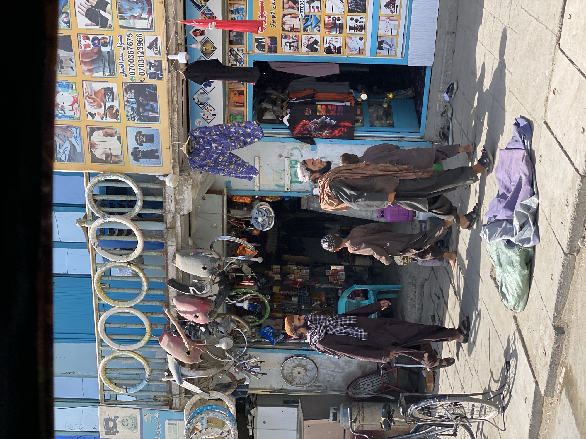 This is a medium shot, captured from a slightly elevated perspective, of a street scene in Kandahar, Afghanistan. The image depicts the front of a shop with a blue facade. Various items are displayed outside and hanging from the shop's exterior. These include several bicycle wheels and what appear to be motorcycle or scooter parts, such as fuel tanks and fenders, suspended from the overhang. A large advertisement poster featuring images of athletic apparel and contact information is affixed to the right side of the shop entrance.
In the foreground, a man wearing a traditional brown shalwar kameez and a checkered scarf walks from left to right. He is accompanied by a young boy, also in traditional clothing, walking behind him. Further to the right, another young boy is visible. A bicycle is parked on the left side of the frame. Piles of fabric are scattered on the sidewalk in the lower part of the image. The environment appears to be a bustling marketplace, with goods displayed for sale and people engaged in daily activities. The lighting suggests it is daytime, with distinct shadows cast on the pavement.