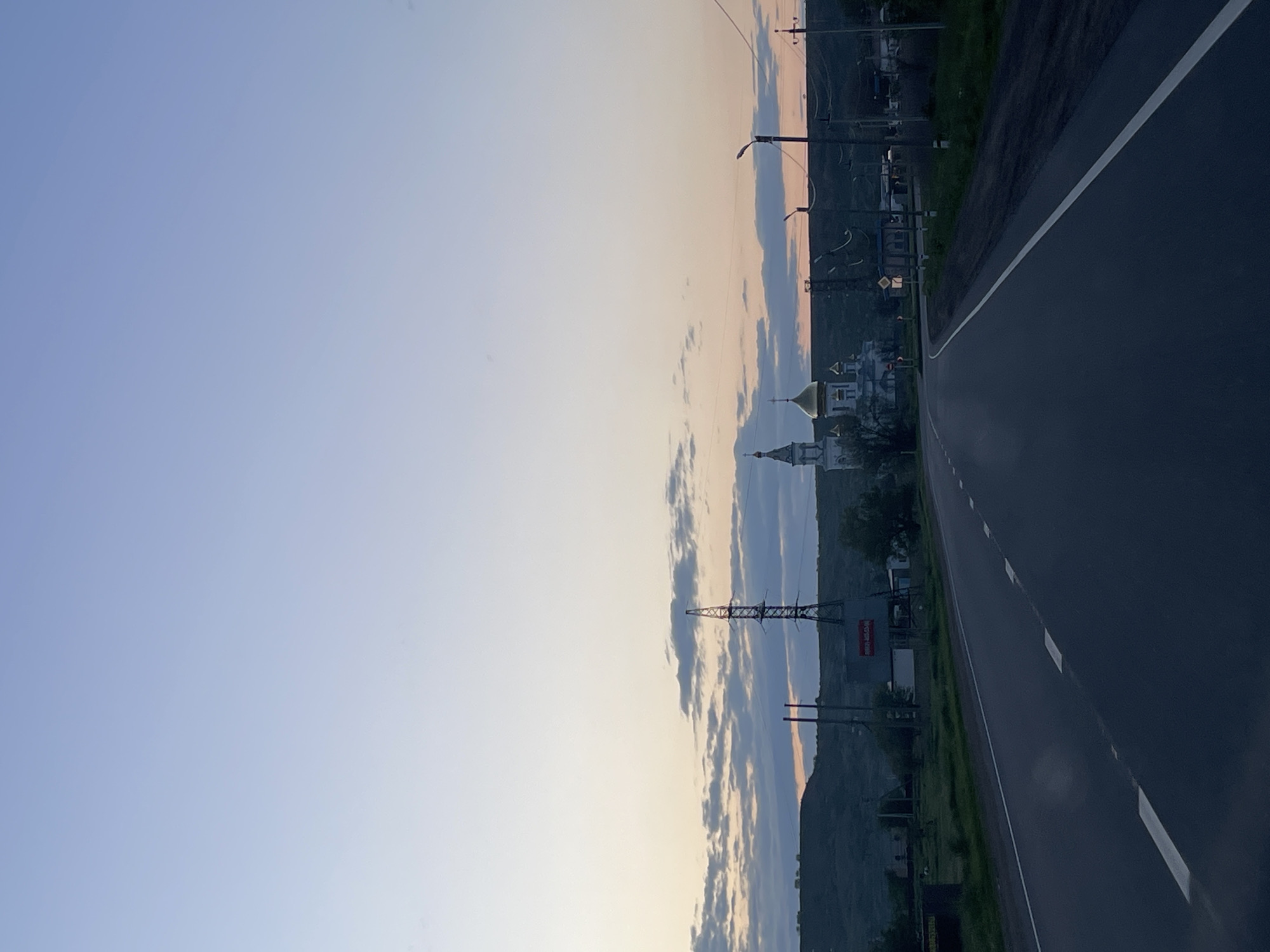 The image depicts a view along a road in the city of Moskovskoye, Russia. The road takes up a large section of the right half of the image, with a white line painted along the edge. A section of land borders the left of the road; atop that land sit several poles and power lines. In the distance along the road, there is a building with a gold dome. The sky is visible above, displaying various shades of blue and orange, suggesting a sunset or sunrise. The overall environment appears to be a mix of urban and rural.