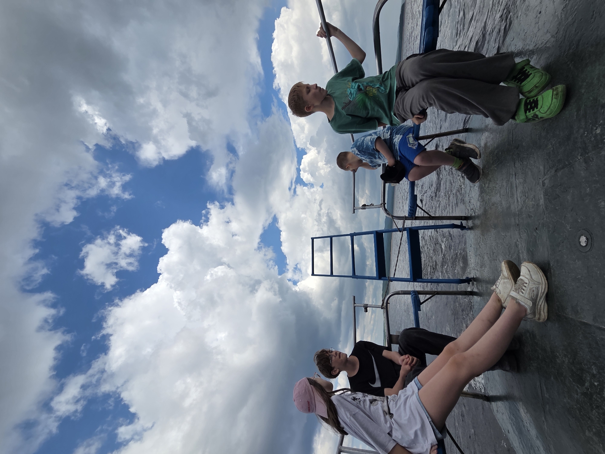 Four children sit on a boat deck on a cloudy day, with a body of water and a distant shoreline visible. The child on the far left, wearing a pink baseball cap and a light-colored t-shirt and shorts, is leaning back. Next to them, a boy with glasses and a black t-shirt sits with his hands clasped. To the right, two other boys are seated. One, wearing a blue tie-dye t-shirt and blue shorts, has a black hat on his lap. The boy on the far right, wearing a green t-shirt and gray pants, is holding onto a metal railing. A blue ladder is positioned behind the first two children. The scene takes place on a boat on the Zeya River in Talakan, Russia.
