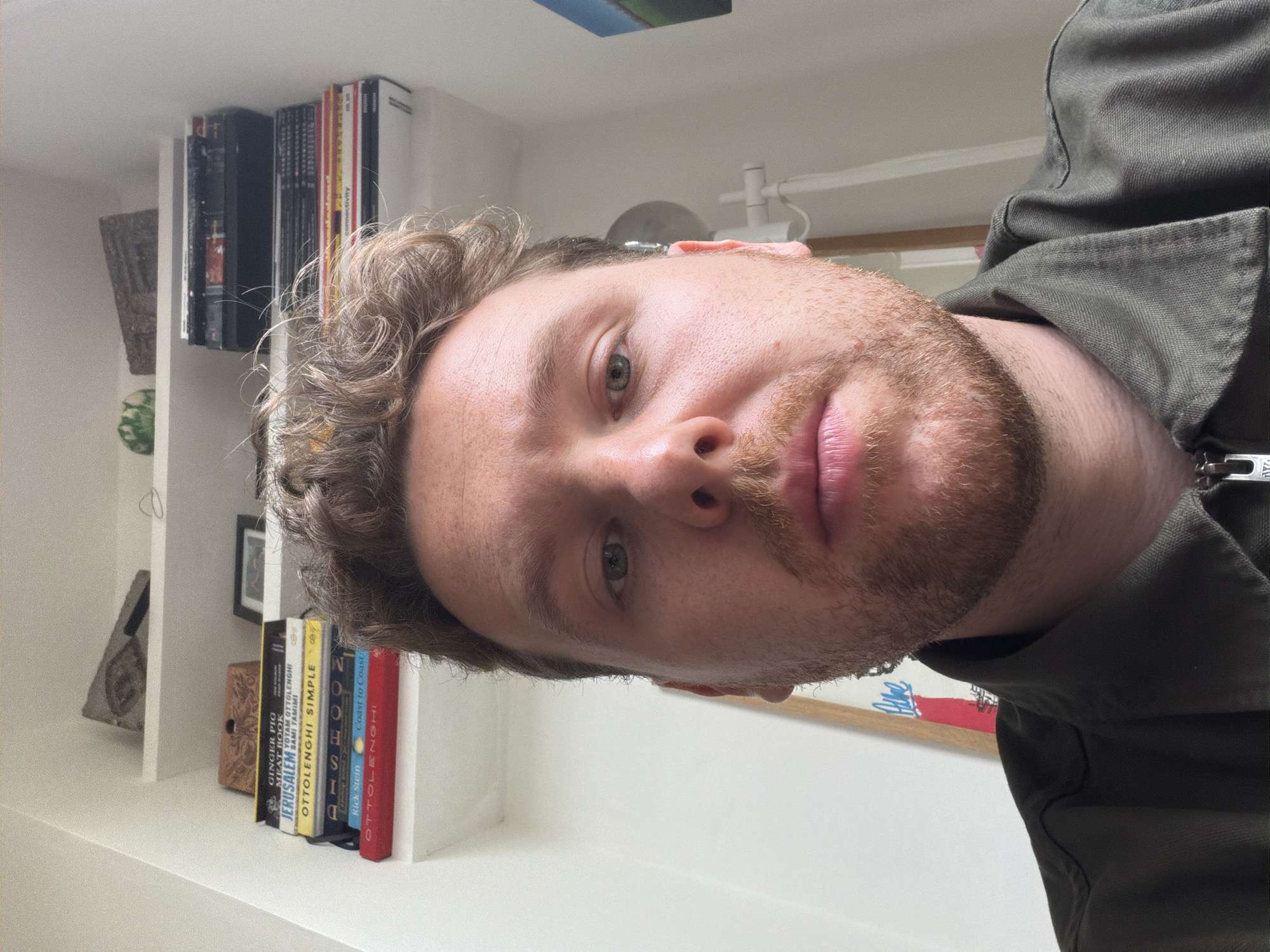 A Caucasian male with curly, light brown hair and a beard is centered in the frame. He is wearing a dark green shirt or jacket. The camera is at an angle, showing him from below. In the background, there is a white shelf with several books, including titles by Ottolenghi (Jerusalem, Simple, Coast to Coast). Other items on the shelf include a woven basket, a framed photo, and a small wooden box. The lighting is soft and natural.