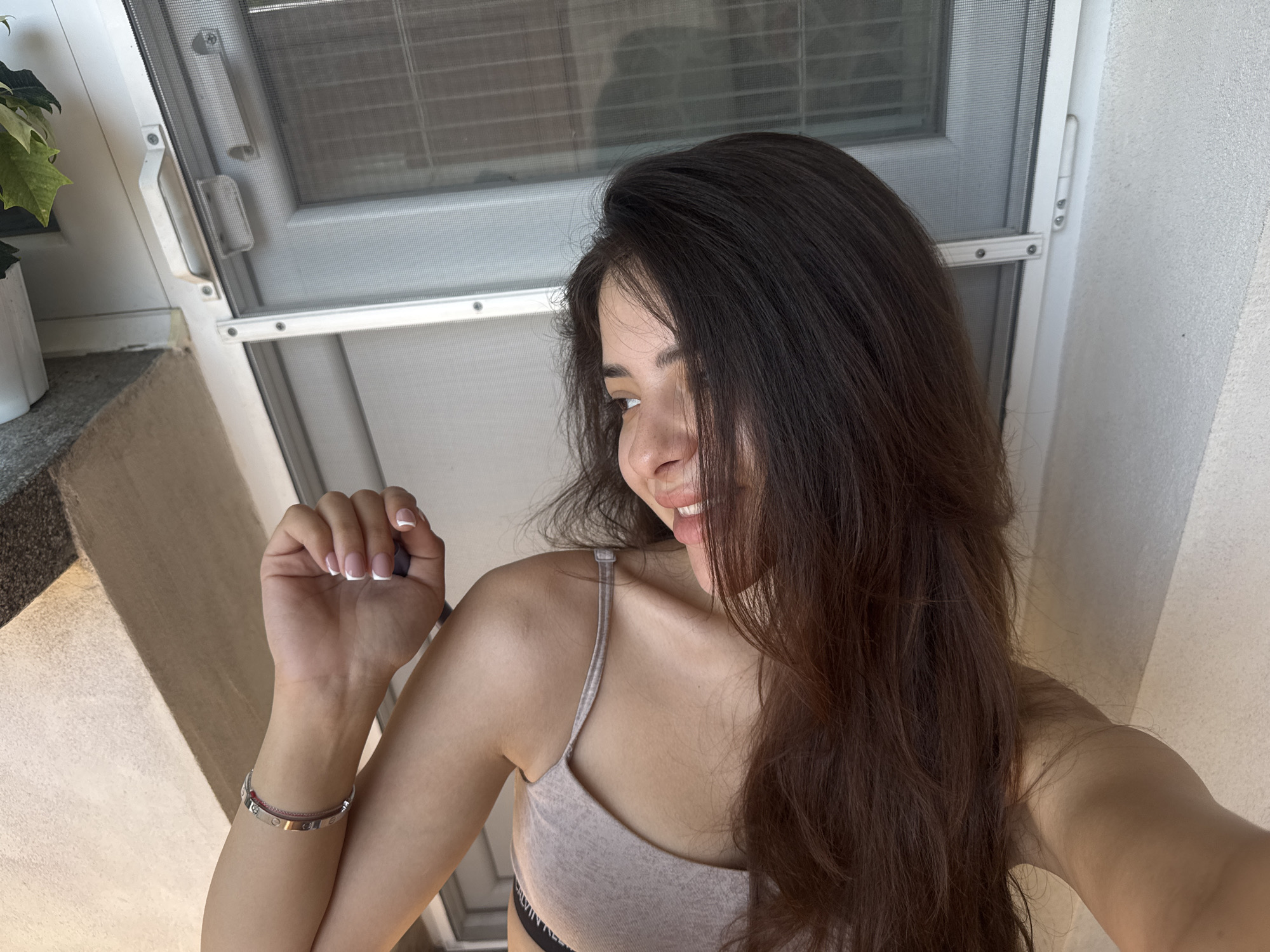 In a selfie taken from a high angle, a young woman with long, dark brown hair and fair skin is captured against a textured off-white wall and a window with a screen. She is wearing a light gray bralette with thin straps. Her left hand, with manicured French-tip nails, is raised near her face, and she is smiling and looking to her left. A silver bracelet with small circular embellishments adorns her left wrist. The background suggests an indoor setting with a glimpse of a plant in a white pot on the left, and the window appears to be a sliding screen door with a white frame and metal handle. The image does not provide specific information to determine its location within the city of Plovdiv, Bulgaria.