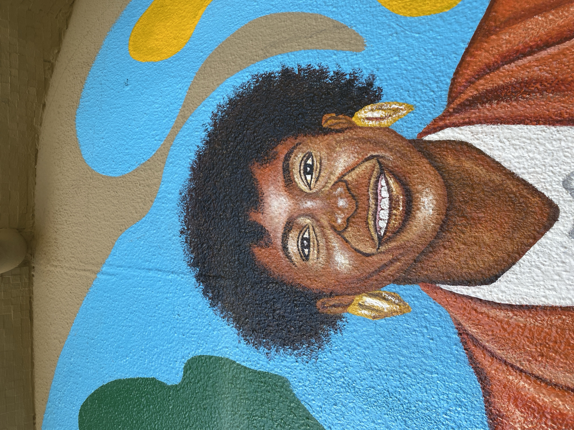 The image shows a close-up of a mural on a wall. The mural depicts a portrait of a smiling Black woman with dark, curly hair and golden earrings. She is wearing a brown garment with a white lining. The background features abstract shapes in blue, green, yellow, and beige. The location, Tawpo, Nigeria, is not discernible from the image.