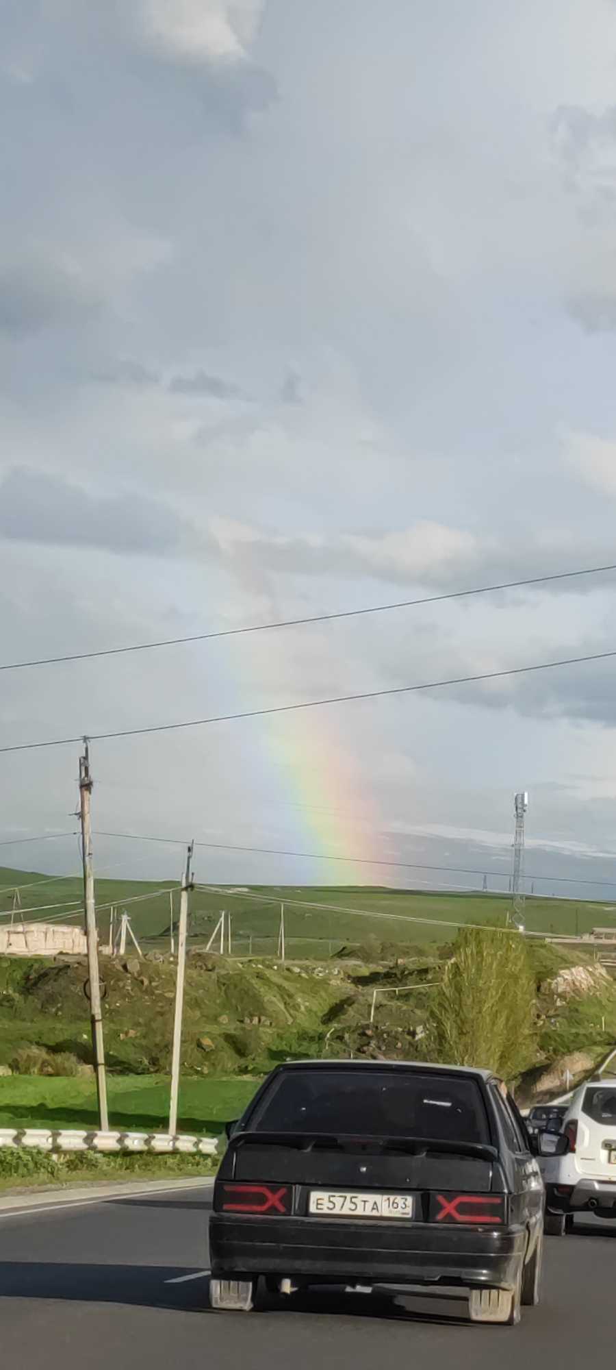 A medium-angle outdoor shot depicts a rainbow in a cloudy sky over a green hilly landscape. Power lines run across the foreground of the image. A black car with license plate E575TA 163 is partially visible in the lower foreground, along with the rear of a white car. The image appears to have been taken from a moving vehicle on a road. Based on the presence of the license plate, the location could be Azatan, Armenia, given the landscape aligns with that region.