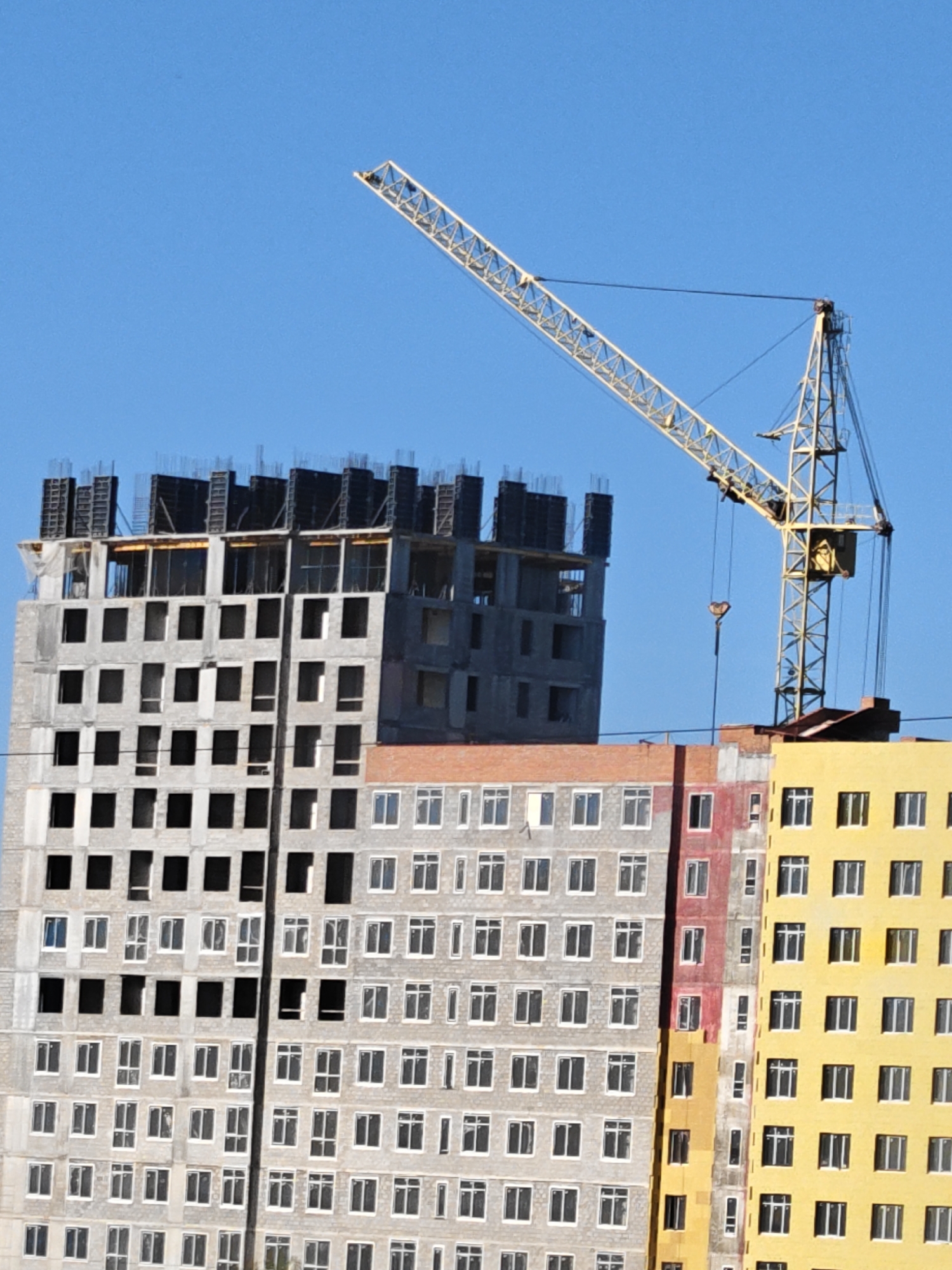 The image shows a construction site in Tobolsk, Russia. Three high-rise buildings are in various stages of completion. The building on the left is a grey concrete structure with many empty window spaces and metal framework visible on its top floors. Adjacent to it is a grey building with installed windows, featuring a red strip on its right side. To its right, a yellow building stands, also with installed windows. A yellow construction crane dominates the upper right of the image, positioned above the buildings. The sky is clear and blue.