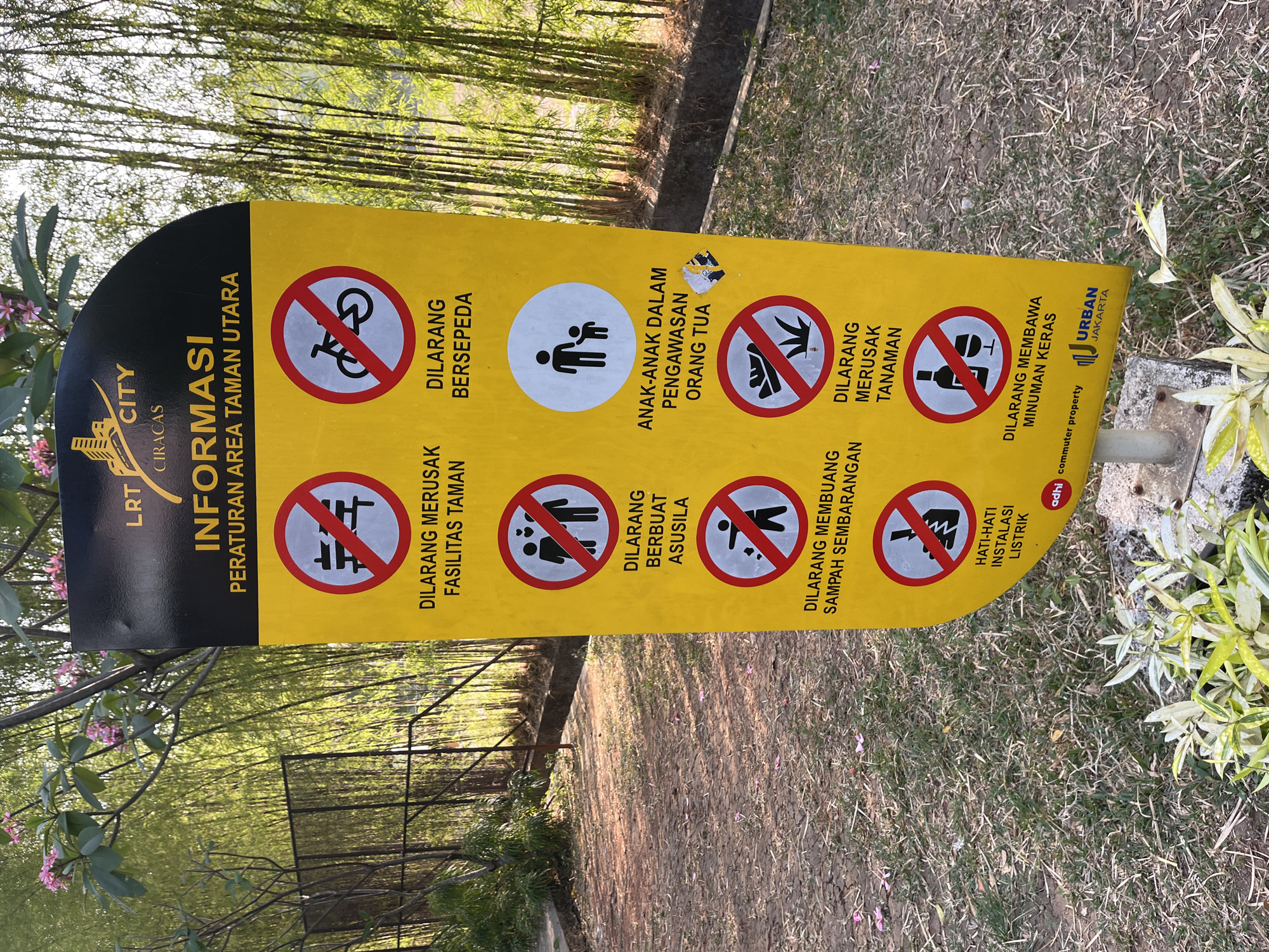 The image shows a yellow information sign with black lettering and icons, located in a park-like setting with bamboo trees in the background. The sign is multilingual, with text in Indonesian and English, and is part of the LRT City Ciracas project in East Jakarta, Indonesia. The sign lists various rules and regulations for the park, including prohibitions against damaging park facilities, littering, inappropriate behavior, and bringing in alcoholic beverages. It also advises children to be supervised by adults and warns about electrical installations. The ground in front of the sign is covered with dry leaves and grass, and there are some small plants in the foreground. The overall impression is that of a well-maintained public space designed to ensure visitor safety and preserve its natural beauty.