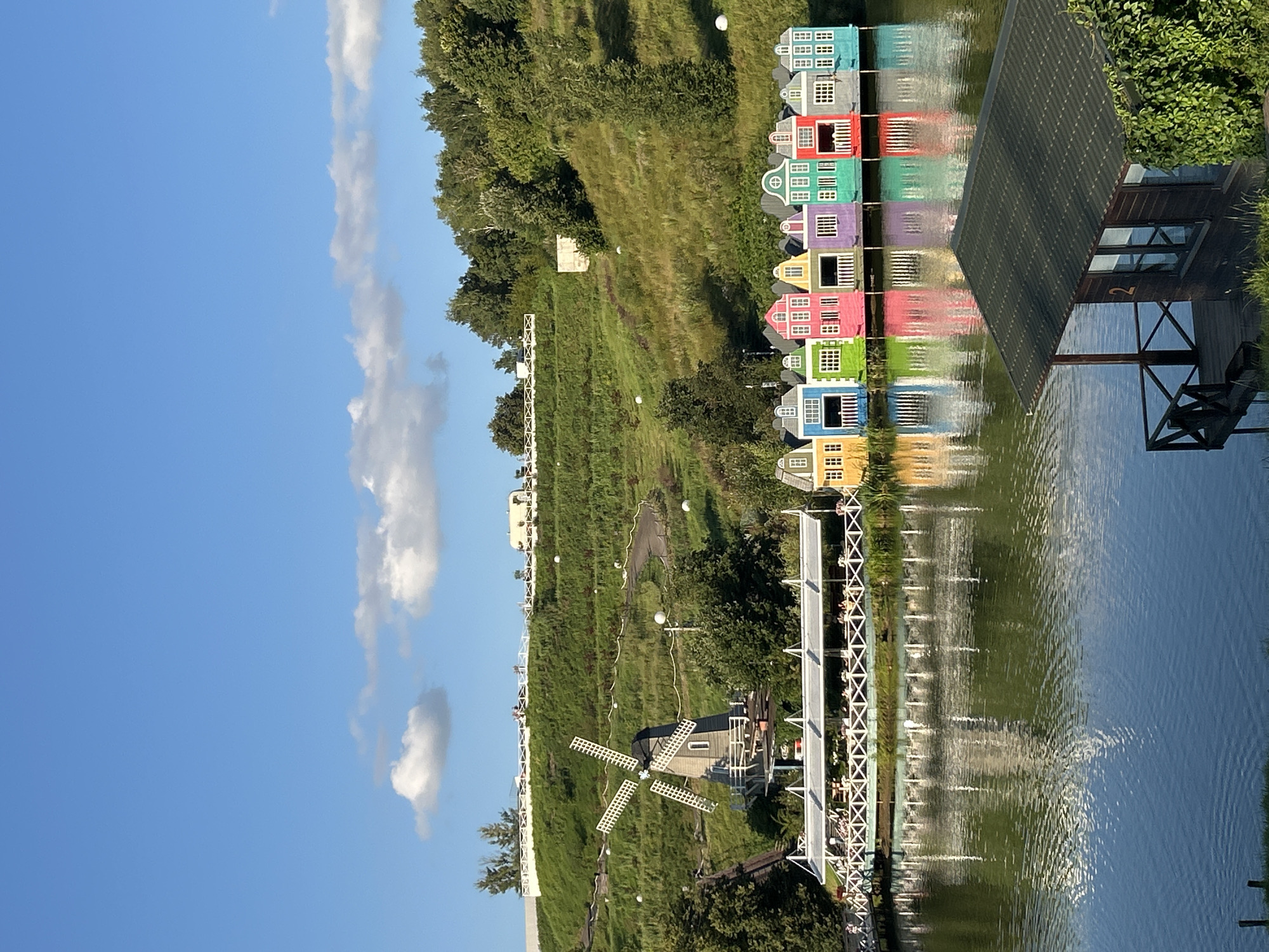A medium shot captures a serene waterfront scene in Romashkovo, Russia, on a bright, clear day. The foreground features the dark-tiled roof and wooden structure of a boathouse, with a visible number '2' on its window, situated on the tranquil, reflective surface of a body of water. In the middle ground, a row of brightly colored, miniature houses, reminiscent of Dutch architecture, are lined up along the water's edge, each with a unique vibrant hue. These houses are connected by a wooden walkway and are reflected in the water. Beyond the colorful houses, a white pedestrian bridge crosses a section of the water, with several individuals visible on it, enjoying the view. To the left, a quaint wooden windmill stands against a backdrop of a lush, green, terraced hillside. The hillside is dotted with white spherical decorative elements and winding pathways, suggesting a park or resort. The upper portion of the image is dominated by a vast, clear blue sky with a few scattered white clouds. The overall impression is one of a picturesque, recreational landscape, likely a tourist destination.