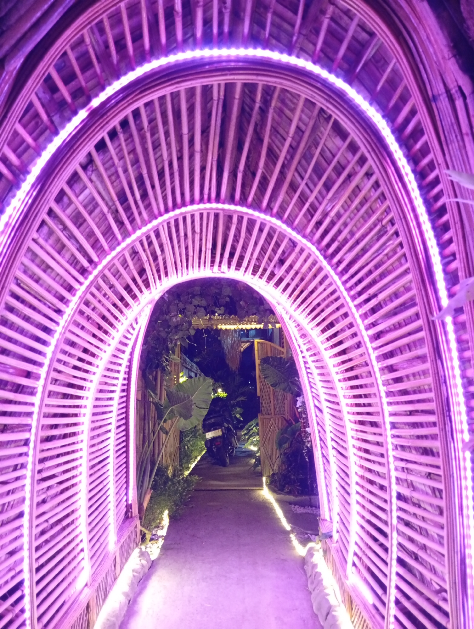 The image shows a pathway leading through a bamboo archway decorated with purple LED lights. The archway is made of intricately woven bamboo slats and curves overhead, creating a tunnel-like effect. The pathway itself is paved and illuminated by small, warm-toned lights along its edges. Beyond the archway, lush green foliage and hints of other bamboo structures are visible, suggesting an outdoor setting like a garden or a resort. A black motorcycle is parked on the right side of the pathway, further into the scene. The overall ambiance is festive and inviting, likely a decorative entrance or walkway in a tropical location. The scene is located in Tampaksiring, Indonesia.