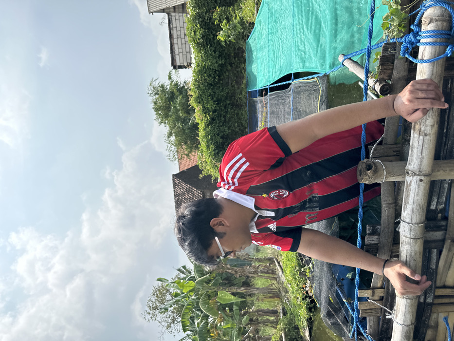 A young person wearing a red and black striped AC Milan jersey and glasses stands next to a bamboo structure secured with blue rope. Their hands are resting on the bamboo. Behind them is a pond with a green tarp and some greenery. The sky is partly cloudy. The image appears to be taken outdoors in a rural setting. Houses and trees are visible in the background.