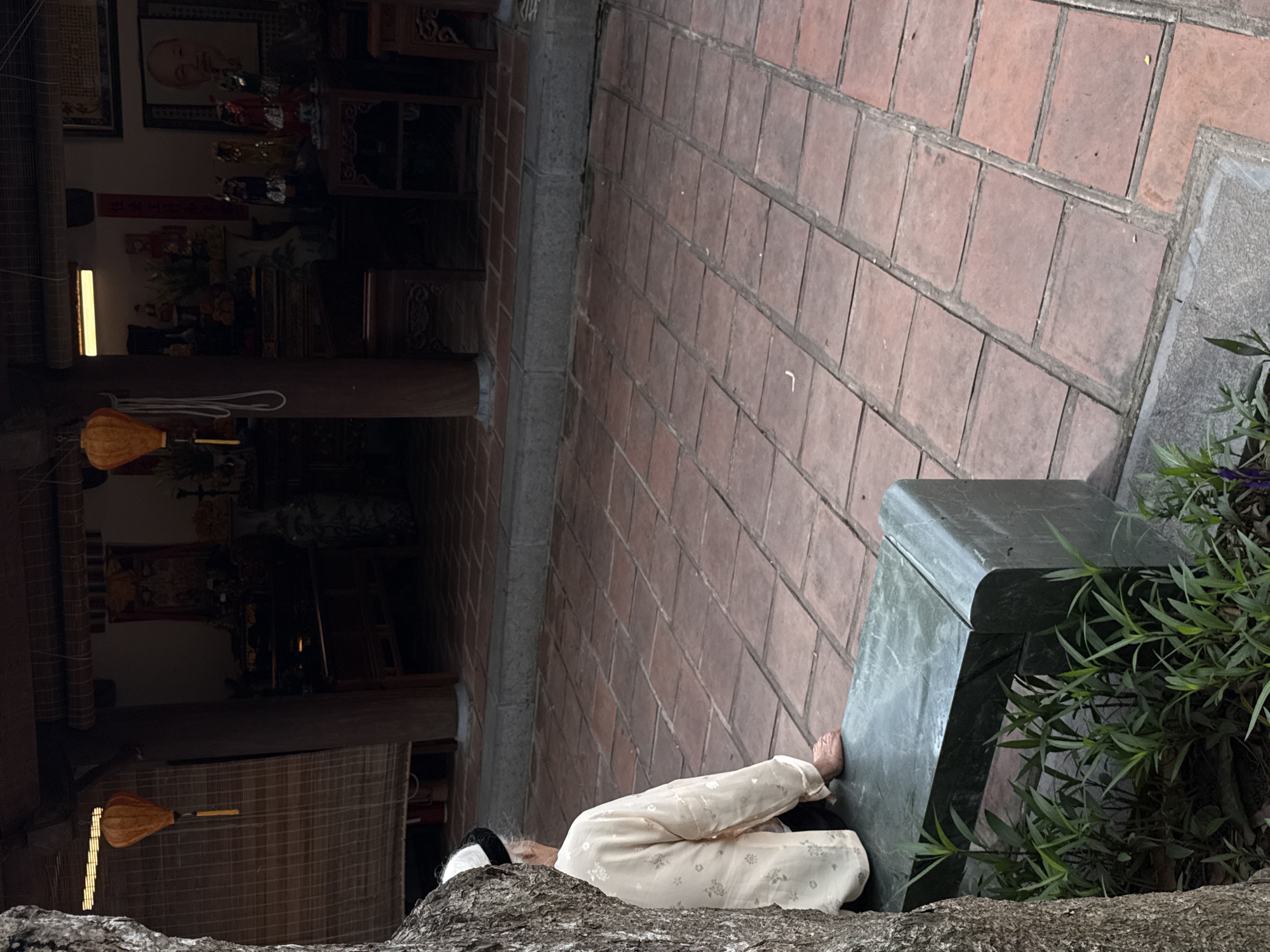 A medium shot depicts a partially visible individual seated on a weathered stone surface in the city of Hà Nội, Vietnam. The person wears light yellow pants with a subtle floral pattern and is positioned near a dark green, possibly metallic, cube-shaped object and a bush. The foreground features the textured surface of the stone they are sitting on. The background includes a red brick walkway. To the left, a traditional Vietnamese interior is visible, featuring wooden beams, lanterns, and what appears to be a portrait.