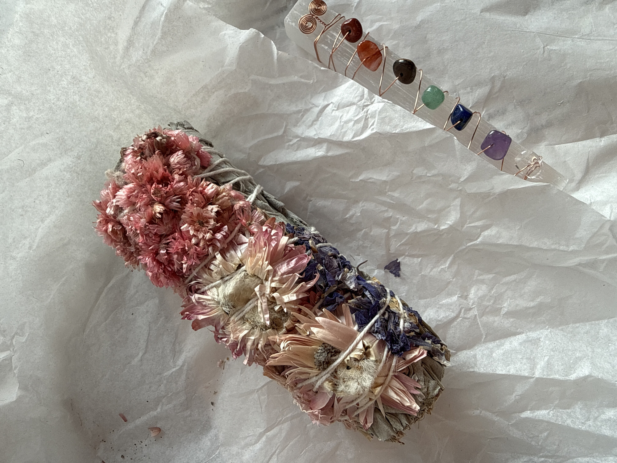The image displays two spiritual or wellness items laid out on a crumpled, off-white, textured paper surface, likely for a product shot or display.
The primary subject in the foreground is a floral smudge stick or bundle, oriented diagonally from the middle left towards the bottom right. It appears to be made of dried sage or similar herbs, tightly bound with white string. Adorning the bundle are several clusters of dried flowers: vibrant pink, small, fluffy flowers at the top-left section, followed by larger, paler pink and cream-colored daisy-like blooms in the middle, and finally, dark purple or blue dried flowers and leaves at the bottom-right section.
In the upper right portion of the image, running diagonally towards the top right, is a slender, translucent white selenite crystal wand. This wand is intricately wrapped with thin, coppery wire that forms small spirals at one end. Along the length of the wand, embedded within the copper wrapping, are seven small, polished, square-cut gemstone beads, each a different color, representing the chakra spectrum: a reddish-orange stone, a darker brown/black stone, a green stone, a deep blue stone, a purple stone, and a clear or very pale stone at the tapered end.
The background consists solely of the crumpled paper, which shows varying shades of light and shadow, indicating bright, possibly natural lighting from an unseen source to the upper left, casting subtle shadows to the lower right of the objects. There is no visible text in the image. The arrangement suggests an aesthetic presentation of items commonly used in cleansing rituals or crystal healing practices.
