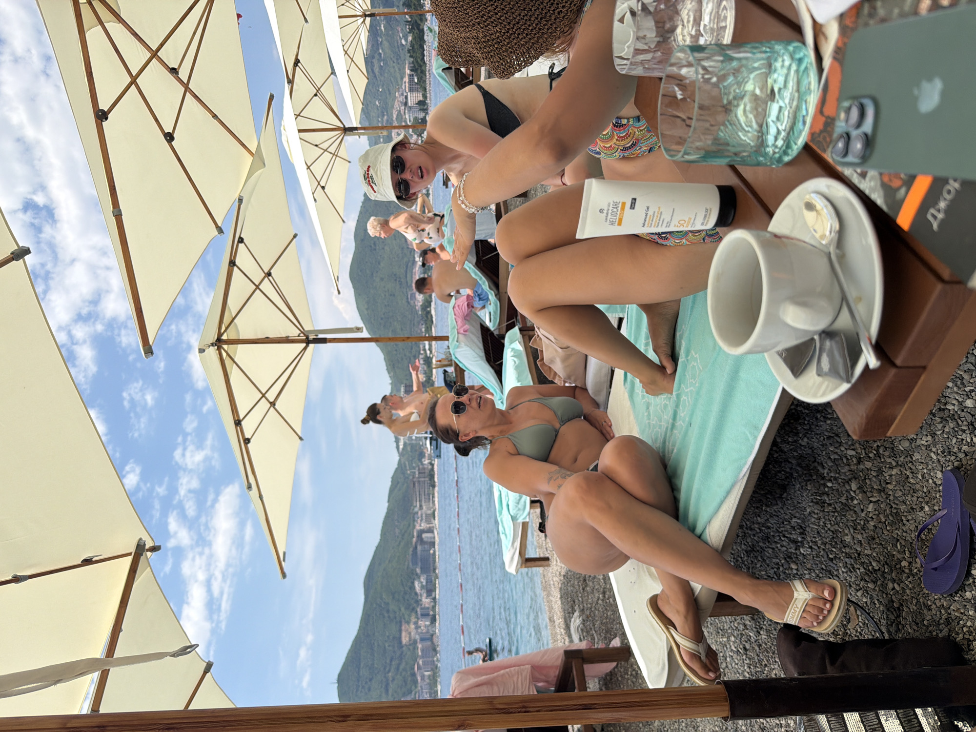 In Budva, Montenegro, this image captures a beach scene under a bright, partly cloudy sky. The foreground features several people relaxing on lounge chairs beneath large, cream-colored umbrellas. One woman in a green bikini is prominently positioned, leaning back on a turquoise towel with her legs crossed. She wears sunglasses and has her arm extended, appearing to gesture or interact with someone out of frame. Another woman, wearing a black bikini and a white bucket hat, is seated to her right, also looking towards the left side of the frame with her hand raised. A white tube of sunscreen, labeled "HELIOCARE," is visible on the table in front of them, along with a white teacup, a spoon, and glasses. The background reveals a body of water, likely the Adriatic Sea, with a town and lush green hills dotted with buildings extending along the coastline. Several other individuals are scattered on the beach and in the water, with one person on a paddleboard in the distance. The beach appears to be covered in pebbles. The overall atmosphere is one of leisure and relaxation on a summer day.
