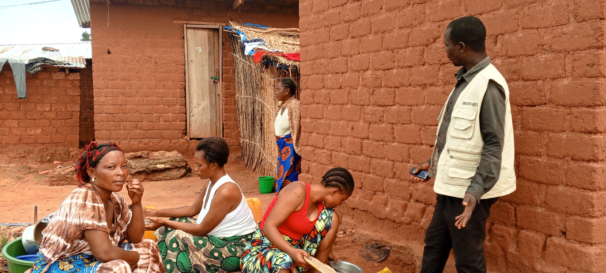 The image captures an everyday scene in Makungu, Democratic Republic of the Congo, featuring six individuals amidst traditional mud brick dwellings. The scene appears to take place outdoors during bright daylight.
In the foreground, three women are seated or crouched on the dirt ground. On the far left, a woman with a red and black headscarf and a brown tie-dye patterned dress looks directly towards the camera, with her hand near her mouth. Beside her, another woman in a white tank top and a green patterned skirt is focused downwards, her hands near a small bowl. To her right, a third woman, wearing a red tank top and a colorful patterned skirt, is crouched, also looking down and engaged with items in front of her, including a metal bowl. A green bucket and some wooden logs are visible on the ground near them.
In the background, a woman wearing a white top and a blue patterned skirt with a light-colored shawl stands near the wooden door of a mud brick house. The door has the number "01" written on it. Adjacent to the door is a structure with a thatched roof, possibly a lean-to or an extension. Further to the left, another mud brick building with a corrugated metal roof is visible, with some clothes draped over it.
On the far right, a man stands, his back partially turned, facing towards the left. He is wearing a dark shirt, black trousers, and a light-colored, multi-pocketed vest. The text "ANOVEVEF3" is visible on his vest. He holds a smartphone in his right hand. The background consists primarily of the textured, reddish-brown mud brick walls of the houses. The overall impression is a communal, domestic setting in a rural environment.