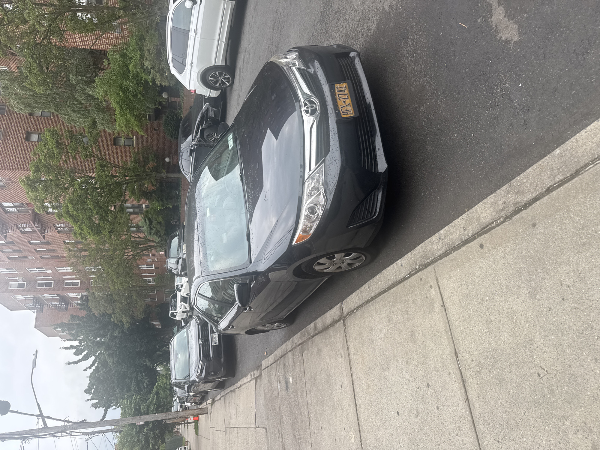 This image depicts a street scene in New York, United States of America. The main subjects are several parked cars along a sidewalk and a brick apartment building. The cars are a mix of colors, including black, white, and silver, and appear to be common passenger vehicles. The building is several stories tall with many windows and some visible air conditioning units. Trees with green foliage are present, suggesting it is daytime during a warmer season. The sky is overcast, and there are visible water droplets on the cars, indicating it might be raining or has recently rained. The sidewalk is made of concrete, and the street is asphalt.
A black Toyota sedan is prominently featured in the foreground, with its license plate clearly visible and readable as "HFX-274Z". Another black car is parked behind it. Further down the street, a white SUV is parked, followed by more dark-colored cars. The general atmosphere is that of a residential urban environment. There are no people visible in the image. The time of day is difficult to determine precisely due to the overcast sky, but the lighting suggests it is likely daytime. The scene is static, with the vehicles parked and no apparent activity taking place.