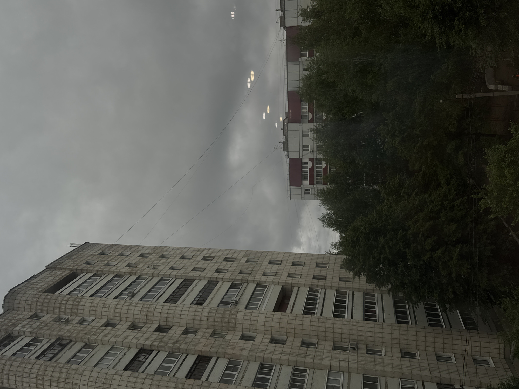 The image presents an urban residential scene in the city of Chaikovsky, Russia, observed under an overcast, uniformly gray sky. The perspective is elevated, likely from a window.
Dominating the left foreground is a tall, multi-story residential building constructed with light gray, rectangular facade panels arranged in a grid pattern. Numerous windows, some with enclosed balconies or loggias, punctuate its surface. Air conditioning units are visible mounted below several windows. The building's architectural style aligns with Soviet-era or post-Soviet residential developments.
A dense line of dark green deciduous trees occupies the midground, largely obscuring the ground level and the bases of the distant structures.
In the midground to the right, a second residential building is partially visible beyond the tree line. This structure features a lighter-colored facade, possibly white, with prominent reddish-brown roof elements or decorative bands. Multiple white-framed windows are discernible.
The sky is entirely overcast, exhibiting a consistent gray tone. Several distinct, bright, circular to oval light reflections are visible against the sky, primarily in the upper right quadrant, suggesting internal lens flare or reflections from the viewing pane. Overhead utility wires stretch across the upper portion of the frame. No human figures or discernible human activity are present in the depicted environment.